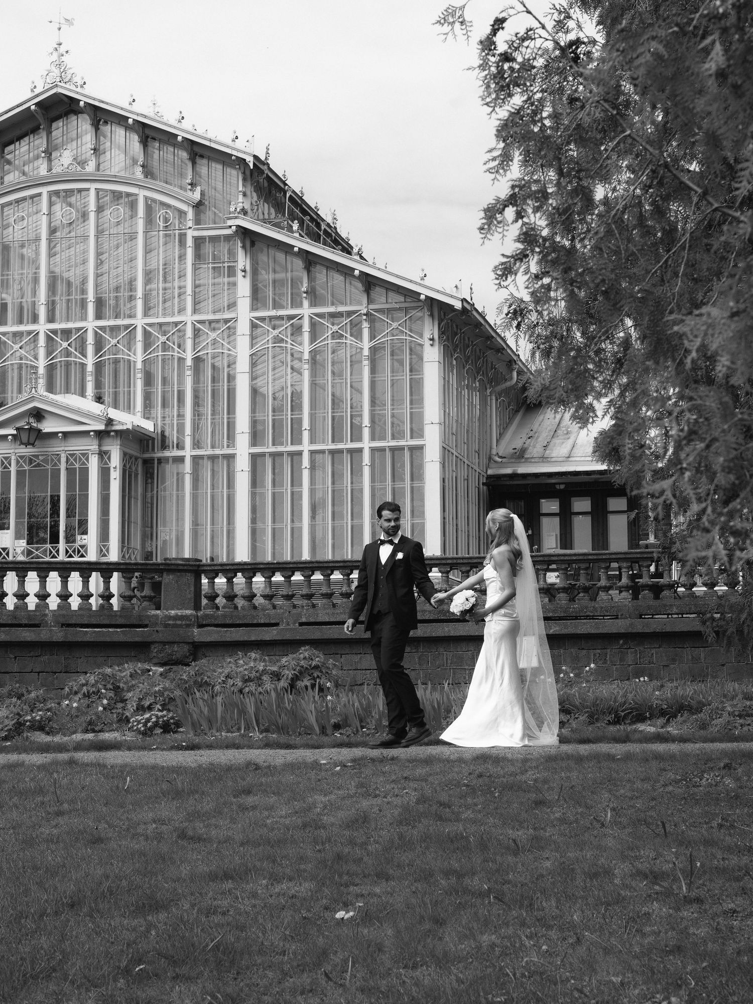 Black and white photo of bride and groom holding hands outside a grand Victorian greenhouse on their wedding day.