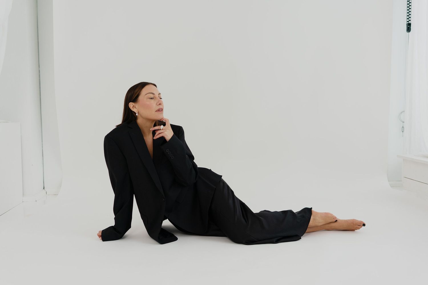 A person in a black outfit reclines on a white floor against a minimalist white backdrop, striking a contemplative pose.