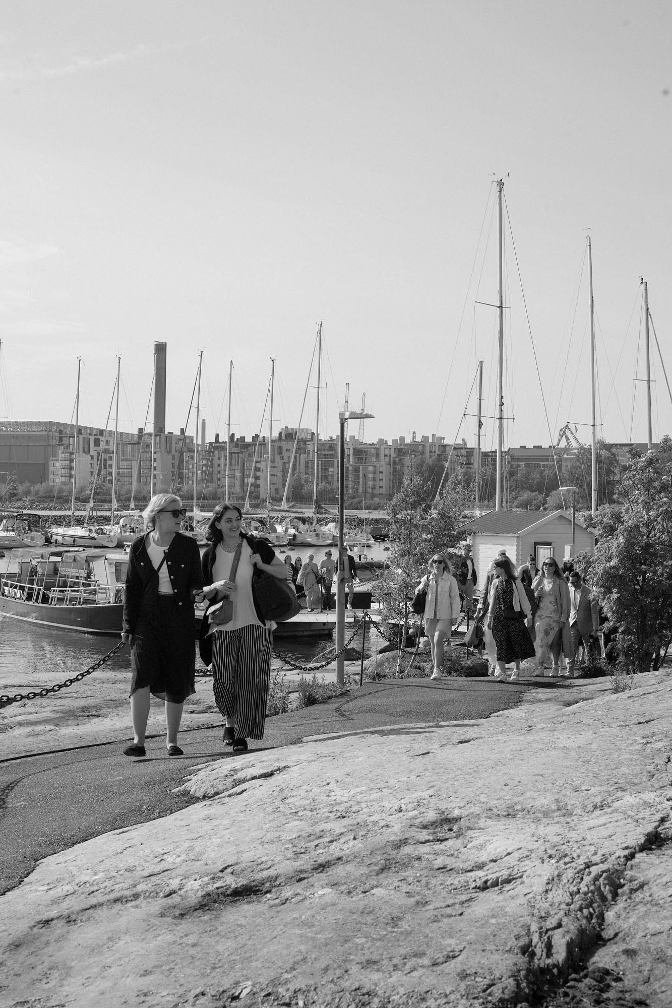 Black and white photo of women walking along a rocky harbor path with sailboats moored in the background.