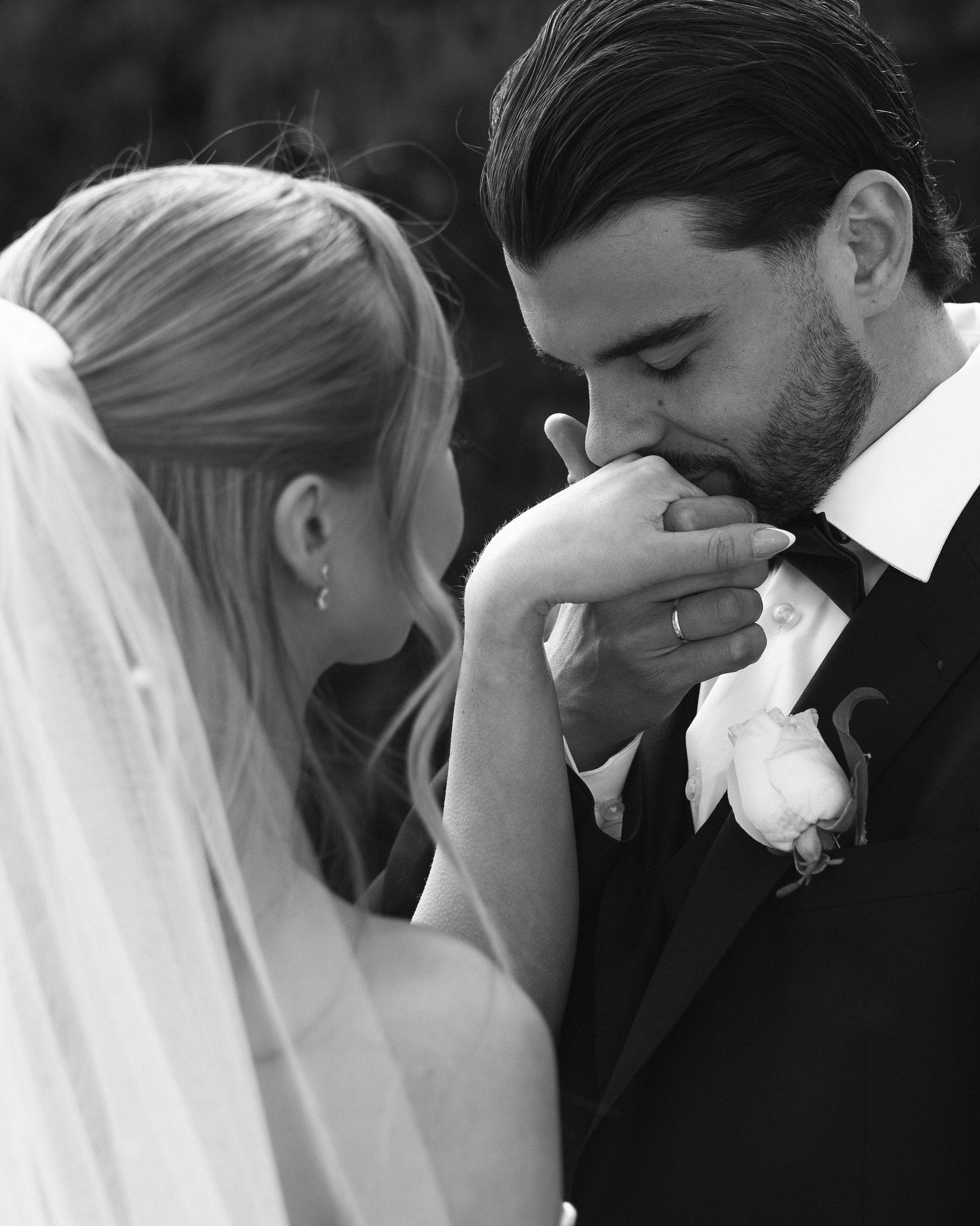 Black and white wedding photo of groom kissing bride's hand, both dressed formally, with veil and boutonniere visible.