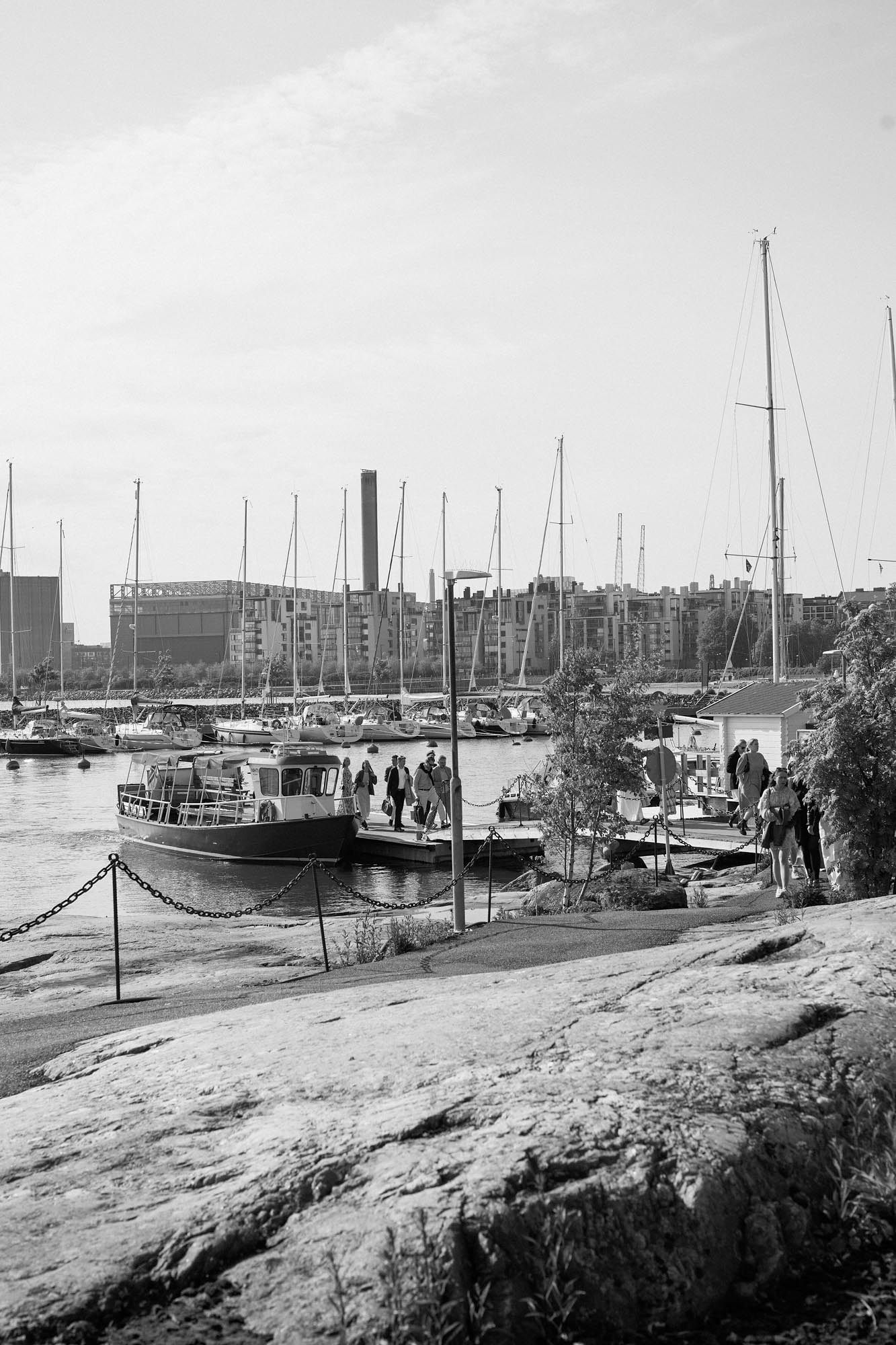 Black and white photo of a marina with sailboats and a motorboat docked near rocky shore, city buildings in background.