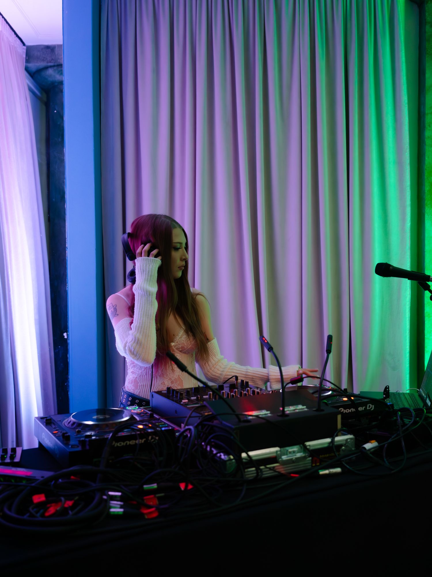 DJ performing at a nightclub with colorful purple and green stage lighting illuminating the booth and backdrop curtain.