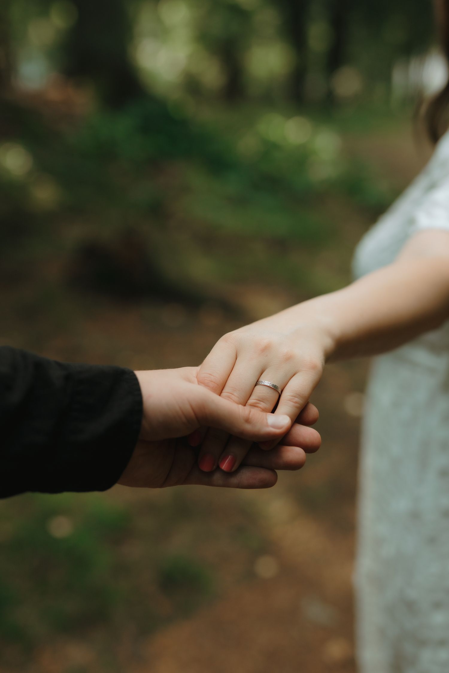Close-up shot of two hands holding each other with gold wedding ring visible against blurred forest background.