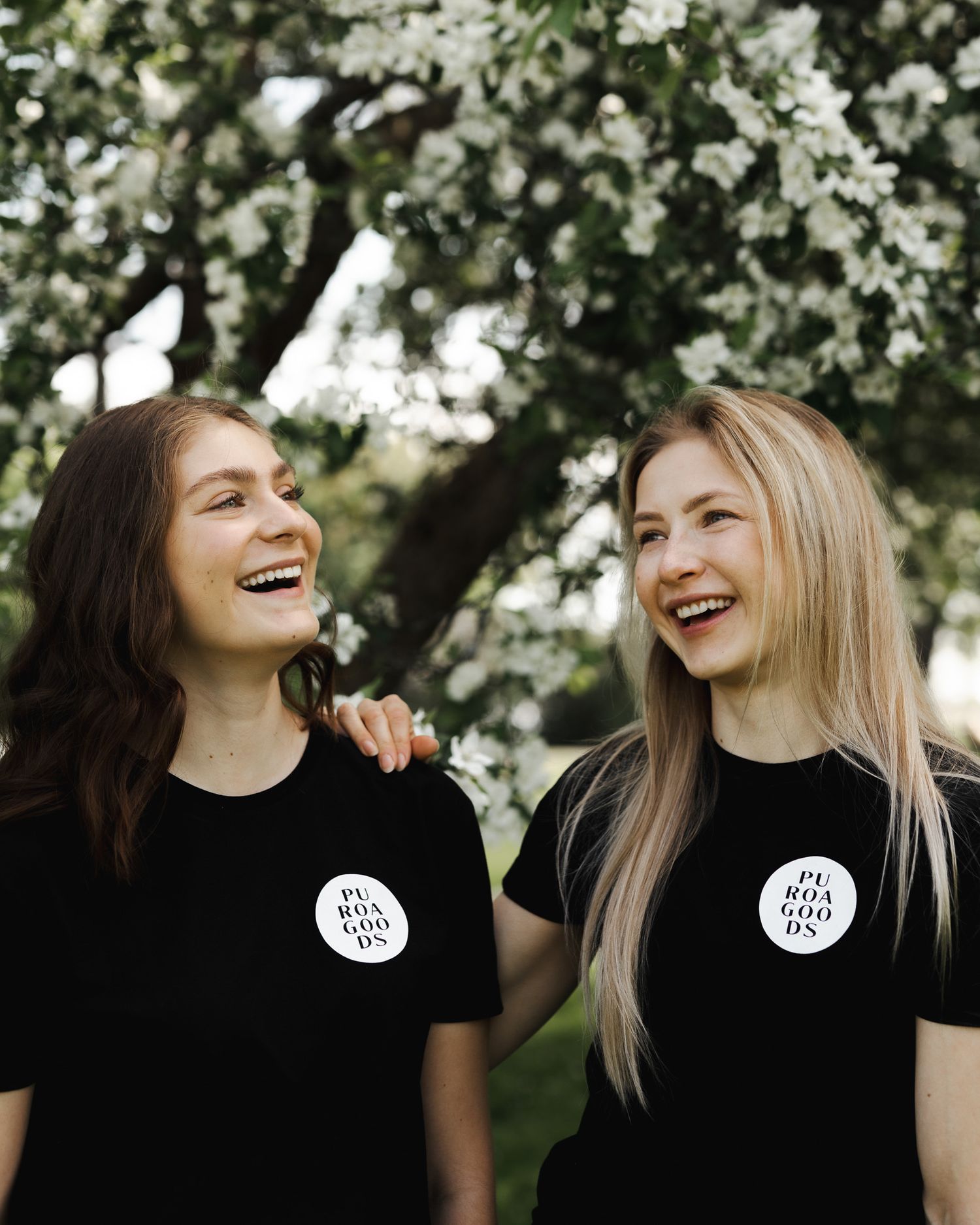 Two friends wearing matching black t-shirts with circular logos share a joyful laugh together under blooming tree branches.