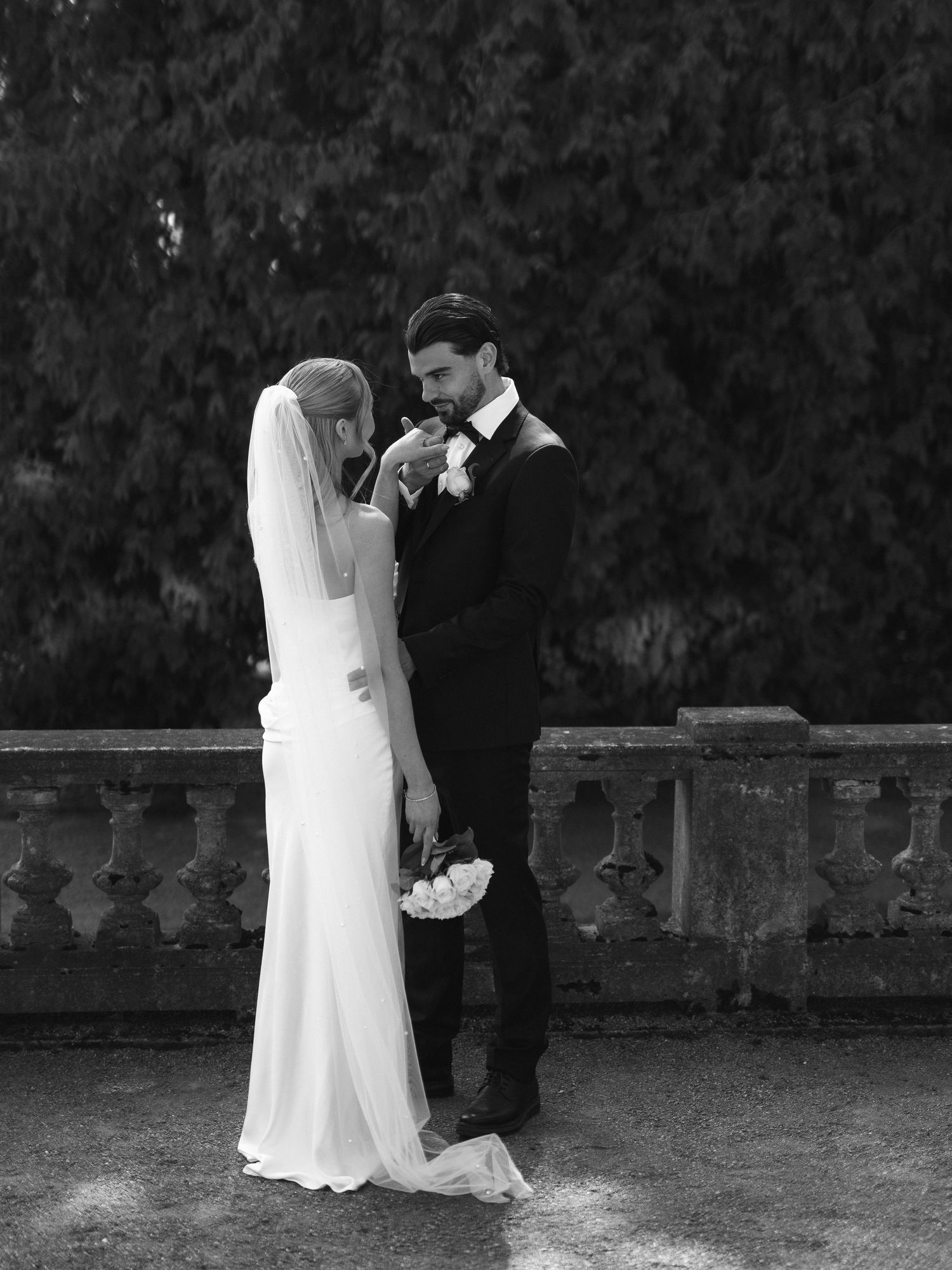 Black and white wedding photo of groom kissing bride's hand on a stone terrace, with lush greenery backdrop.