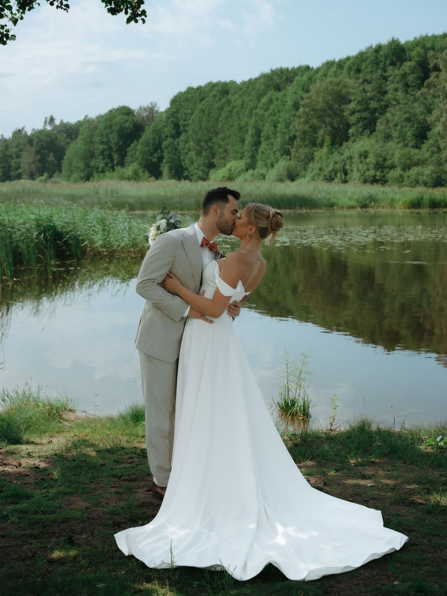 Newlyweds share a romantic kiss by a serene lake with lush green forest backdrop during their summer wedding celebration.