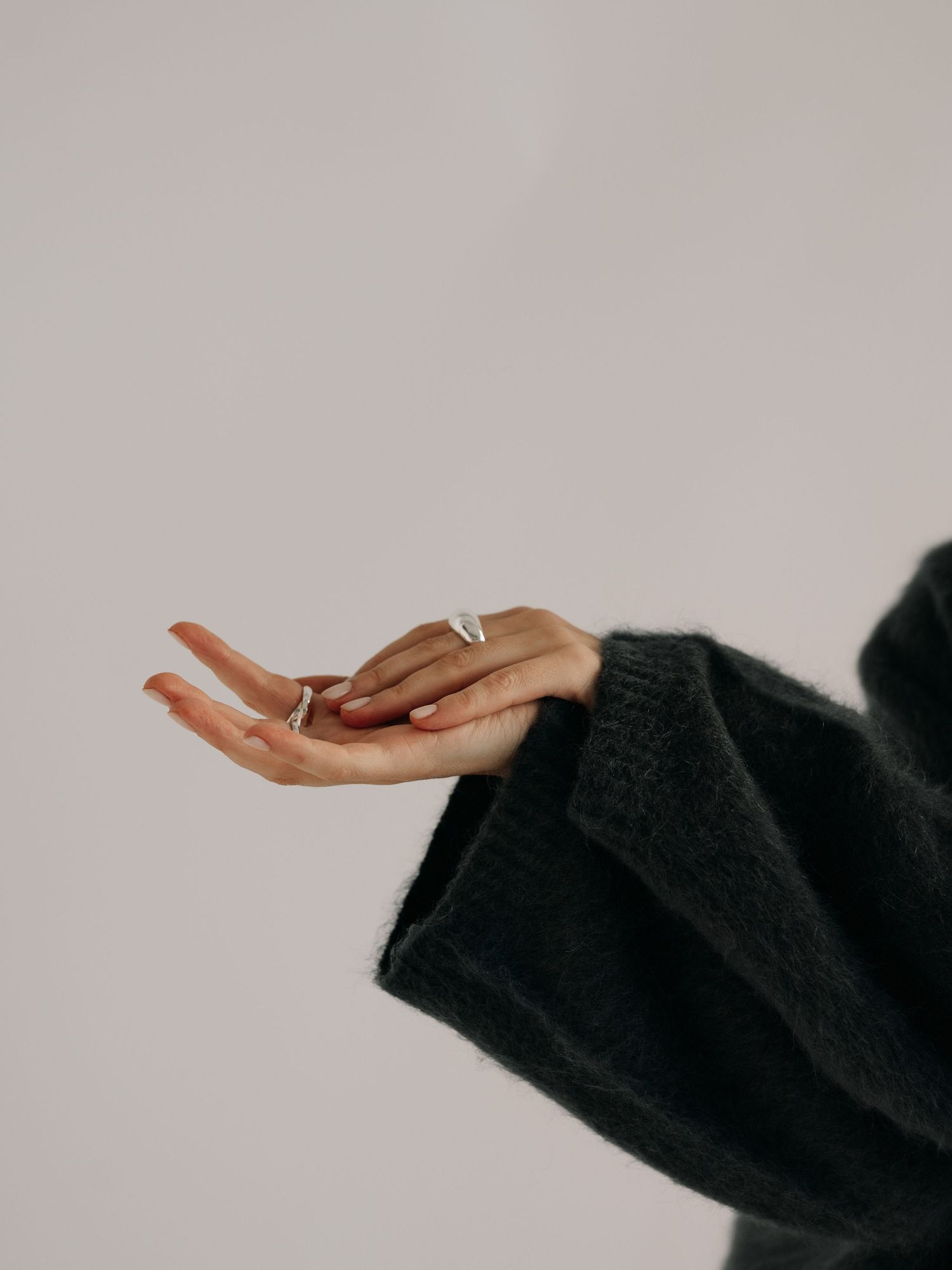 Person wearing a black sweater holds out hand with rings against a neutral grey background.
