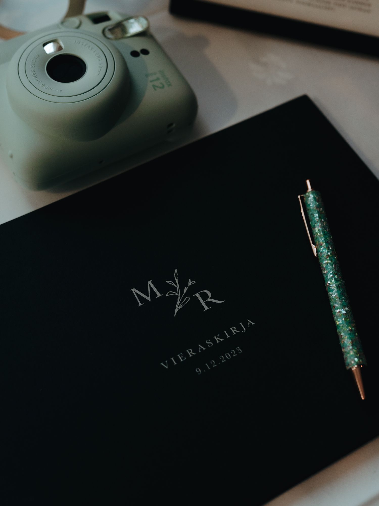 Black wedding guest book with M and R initials next to a mint green instant camera and sparkly pen on a table.