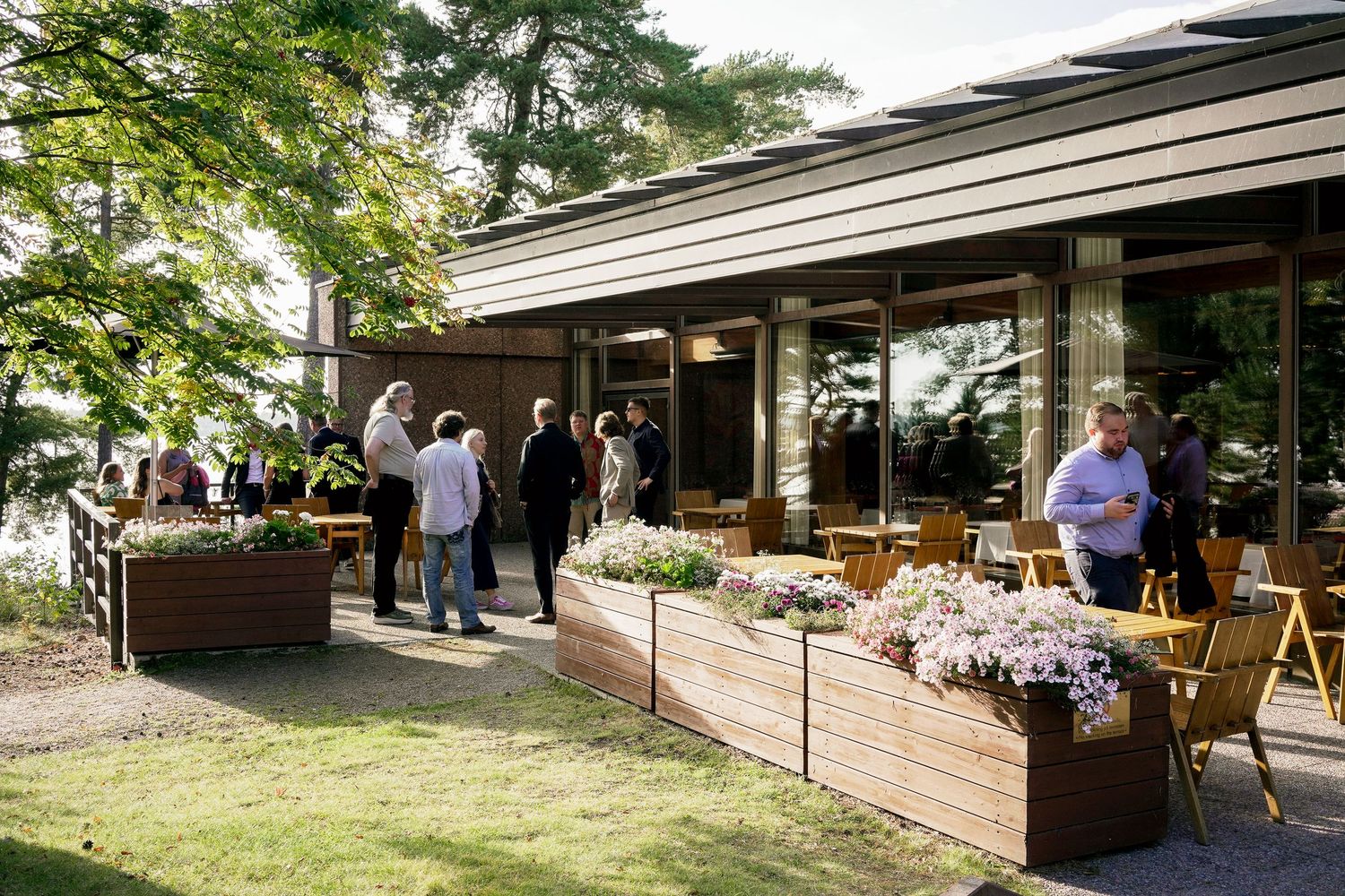 Guests mingle on a modern restaurant patio with wooden planters of flowers, surrounded by trees and large glass windows.