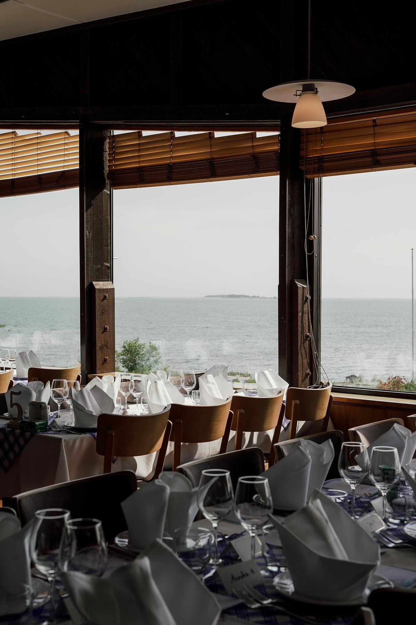 Elegant seaside restaurant dining room with set tables, wine glasses, and panoramic ocean views through large windows.