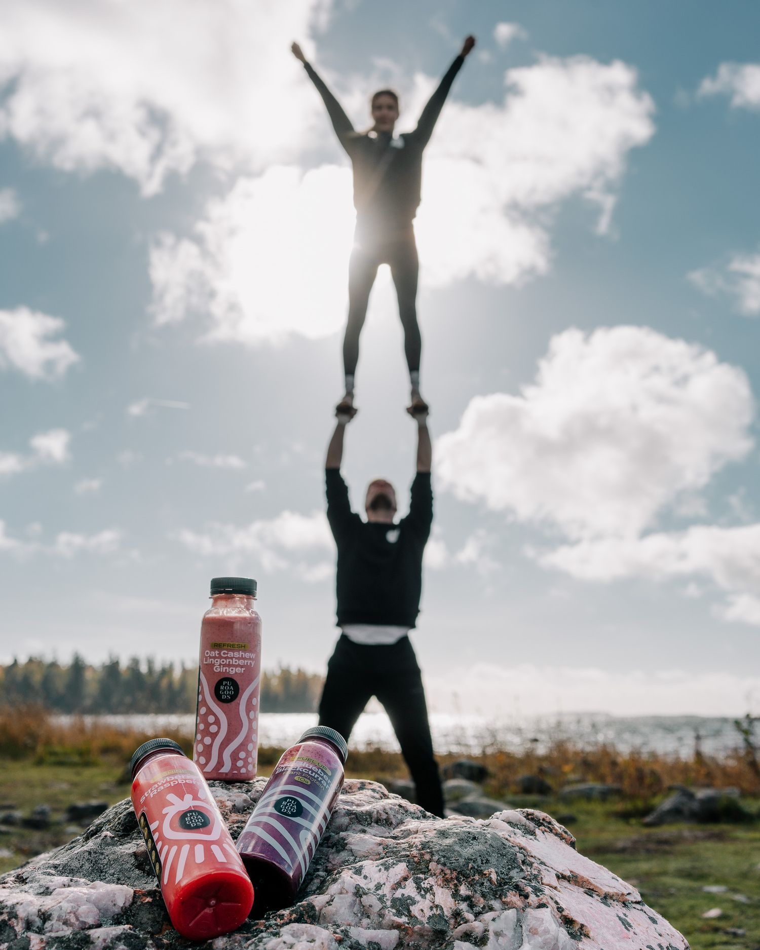 Two people performing an acrobatic lift pose with energy drinks displayed on rocks against a cloudy sky and lake background.