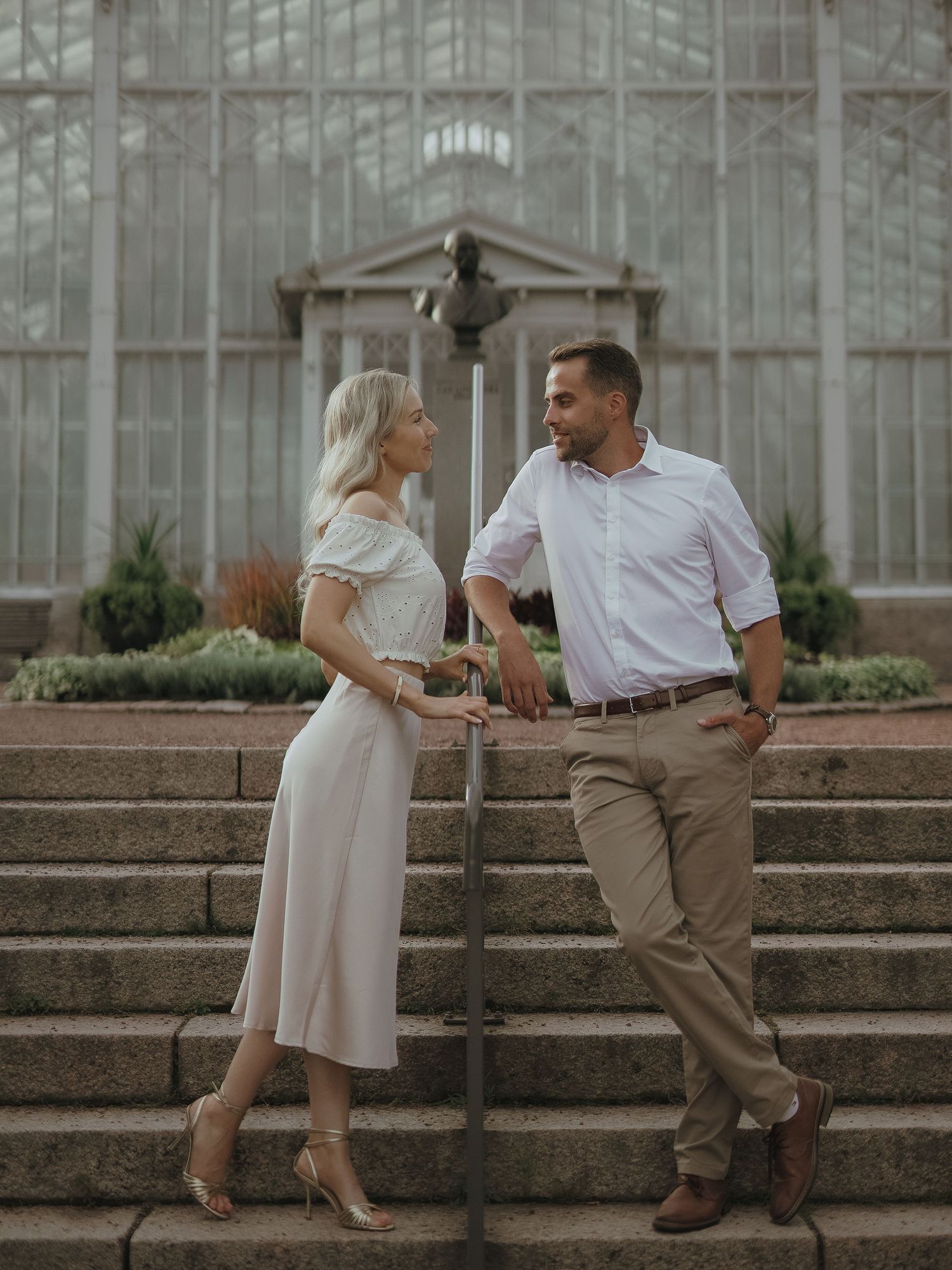 Romantic couple in elegant attire standing on stone steps near a conservatory or greenhouse on a foggy day.