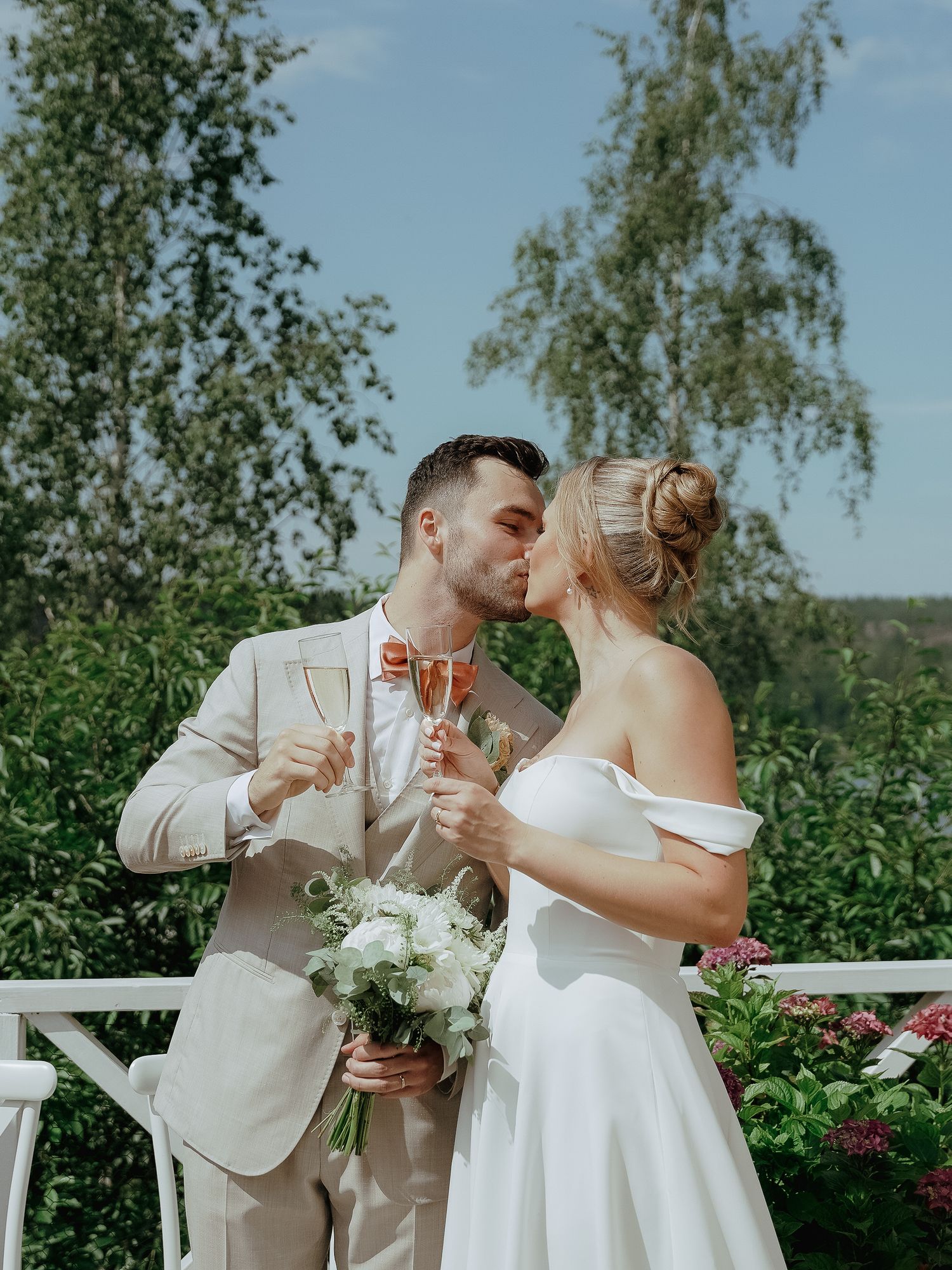 A couple shares a romantic moment in their wedding attire while holding champagne glasses in a lush garden setting.