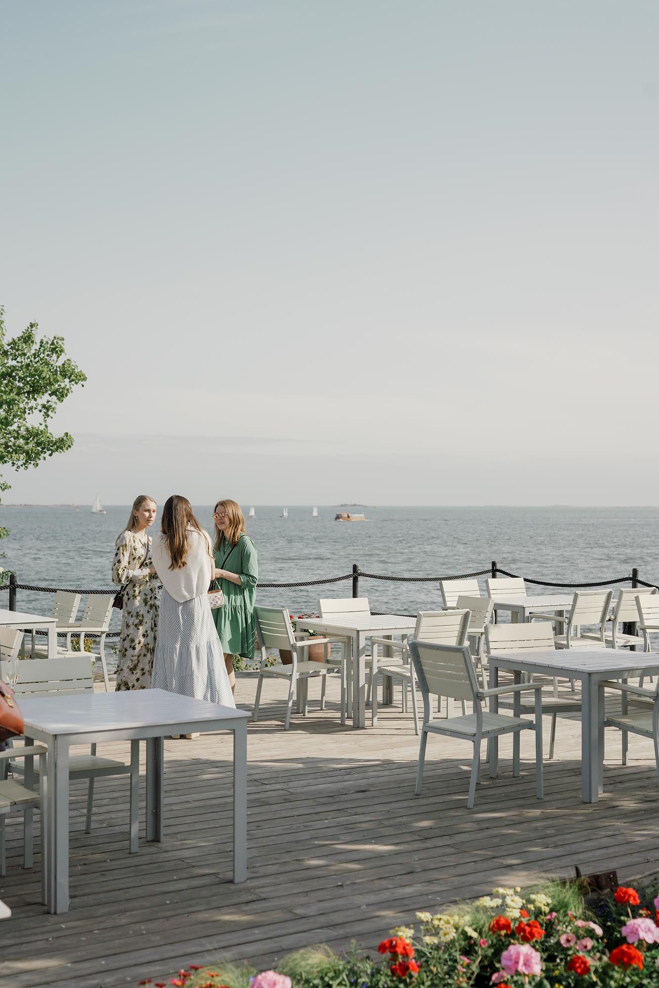 Three women in summer dresses chat on a waterfront restaurant terrace with white tables and ocean views.