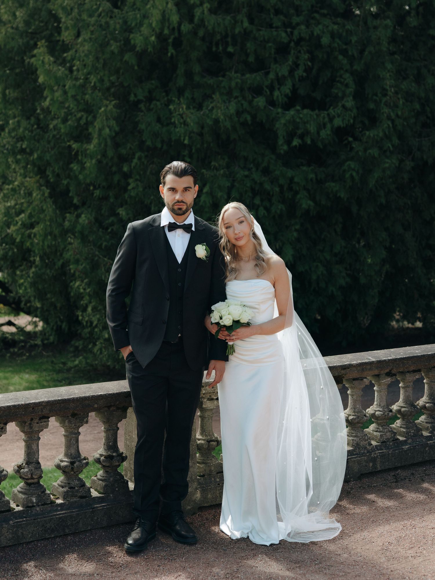 Elegant bride and groom posing on a stone balcony, bride in white gown with veil, groom in black tuxedo with bow tie.