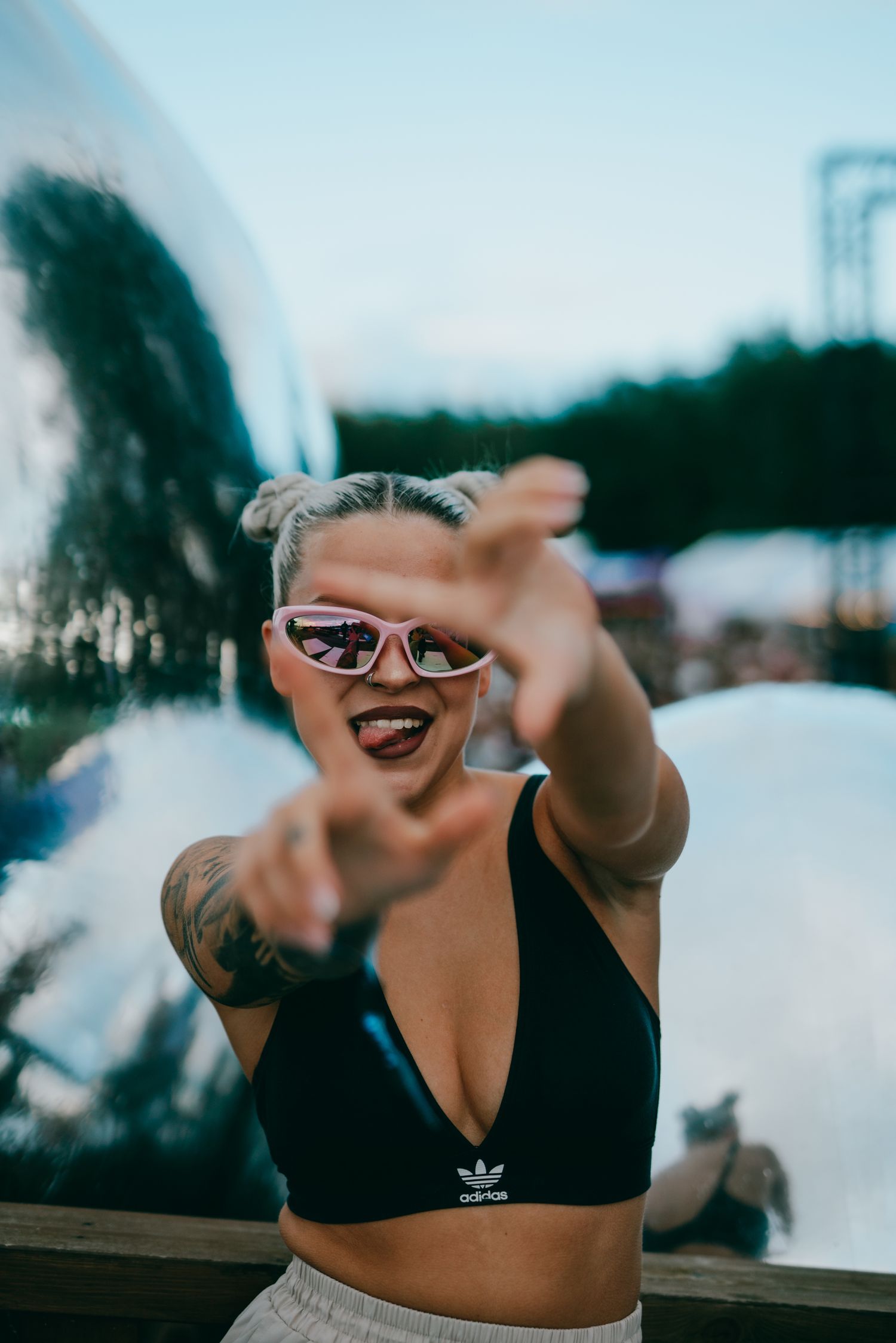 A smiling person in sunglasses and an Adidas sports bra reaching towards the camera with an outdoor background.