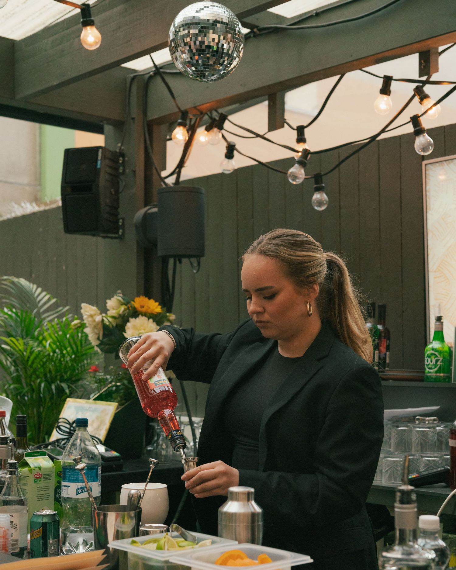 A bartender wearing black pours a drink at an indoor venue with string lights and a disco ball overhead.