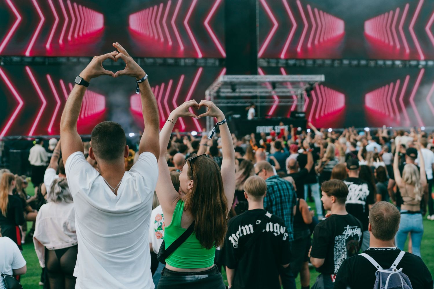 Concert crowd dances with raised arms under neon pink chevron lighting at an indoor electronic music festival.