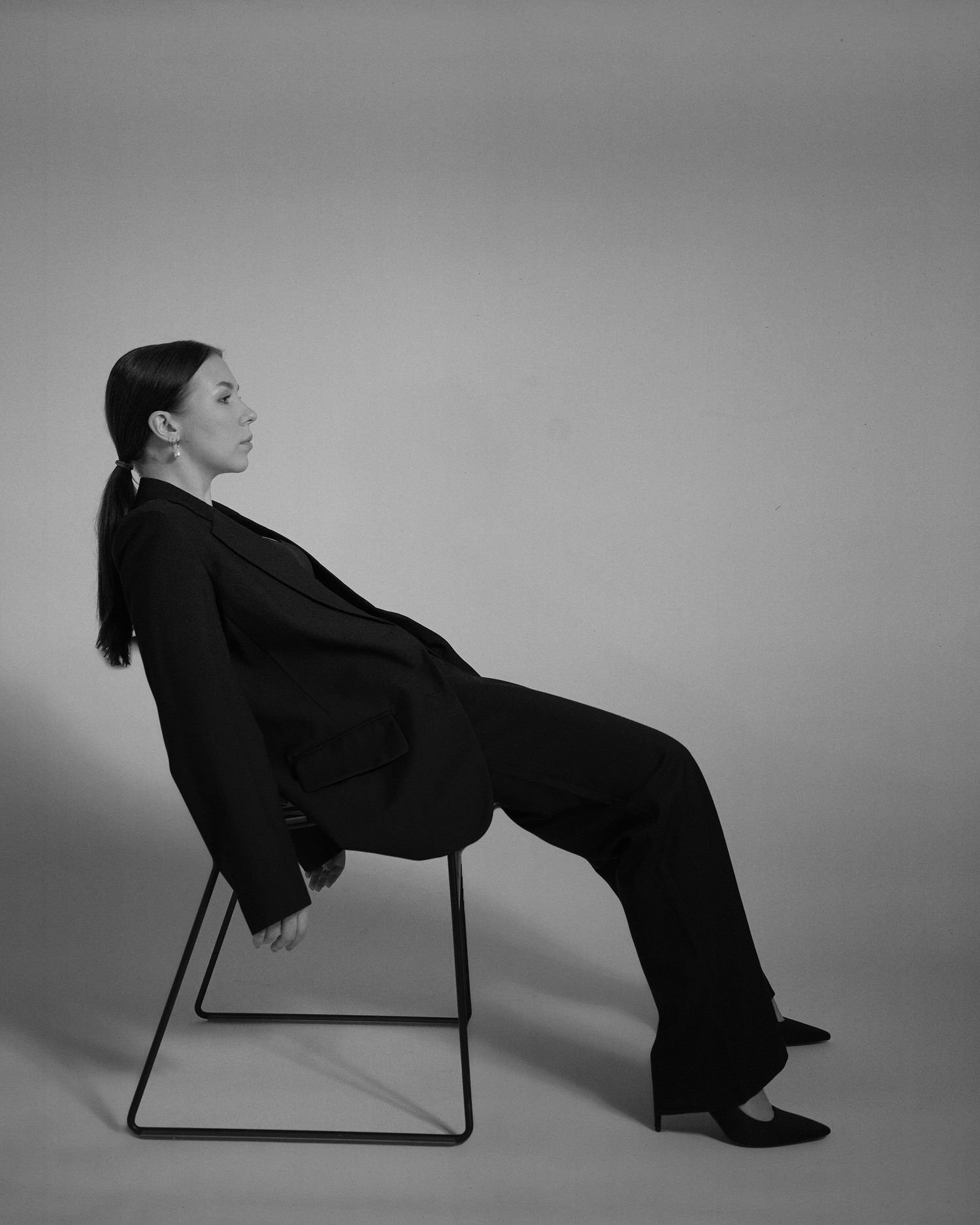 Black and white portrait photograph of a person in a dark suit reclined on a minimalist chair against a plain background.
