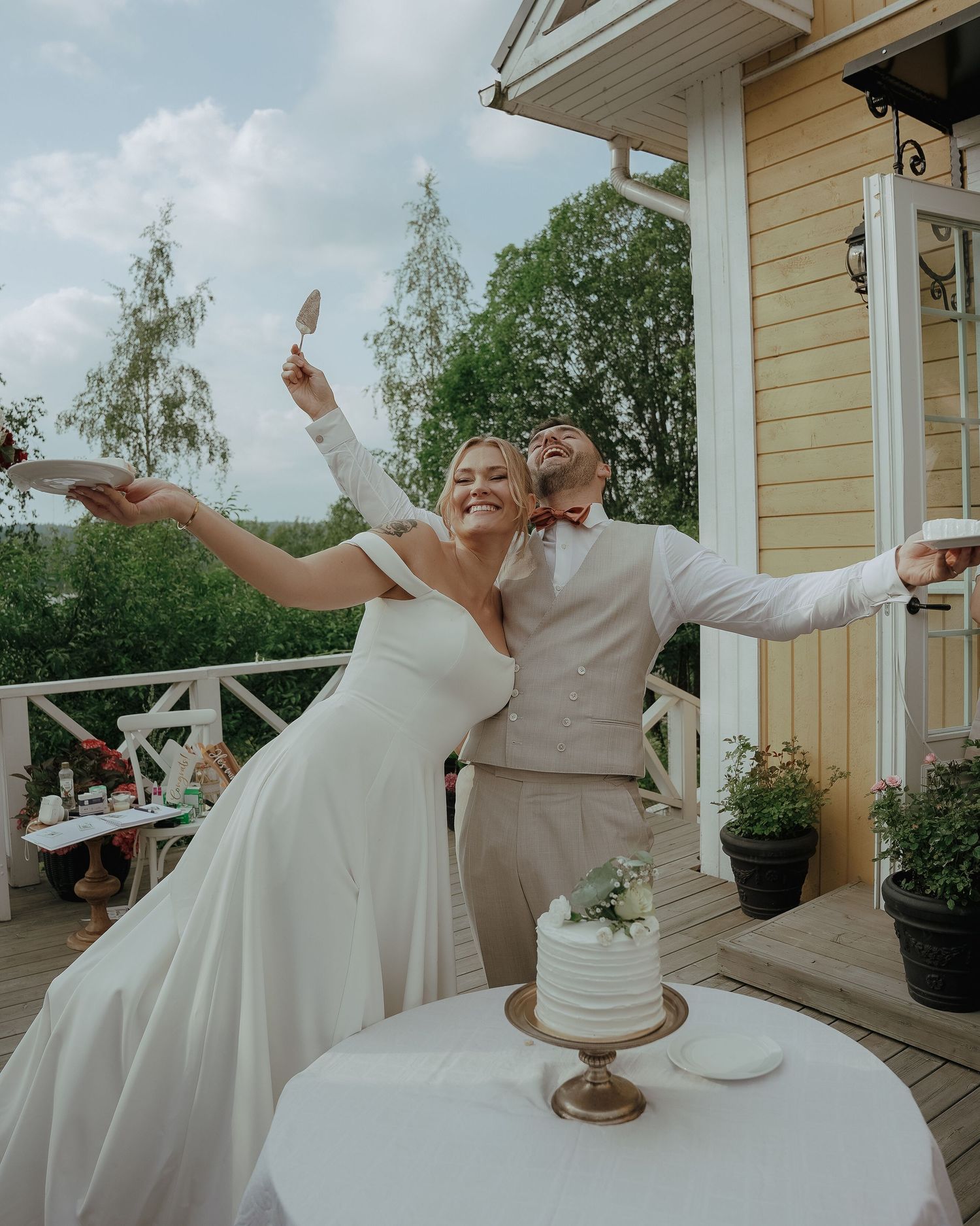 Joyful wedding couple spreading their arms in celebration on a porch next to their white cake.