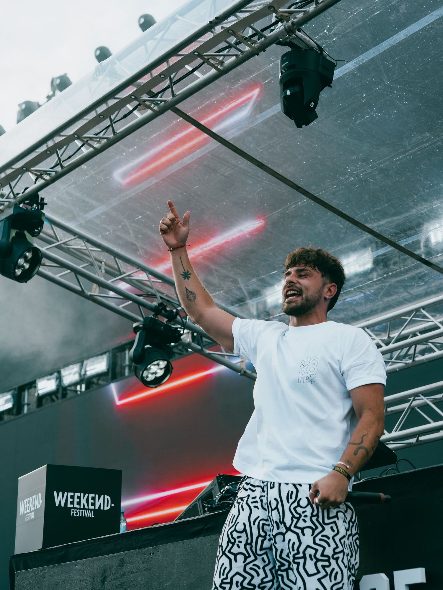An energetic performer in white t-shirt and patterned pants raises their hand on stage under red lighting at Weekend festival.