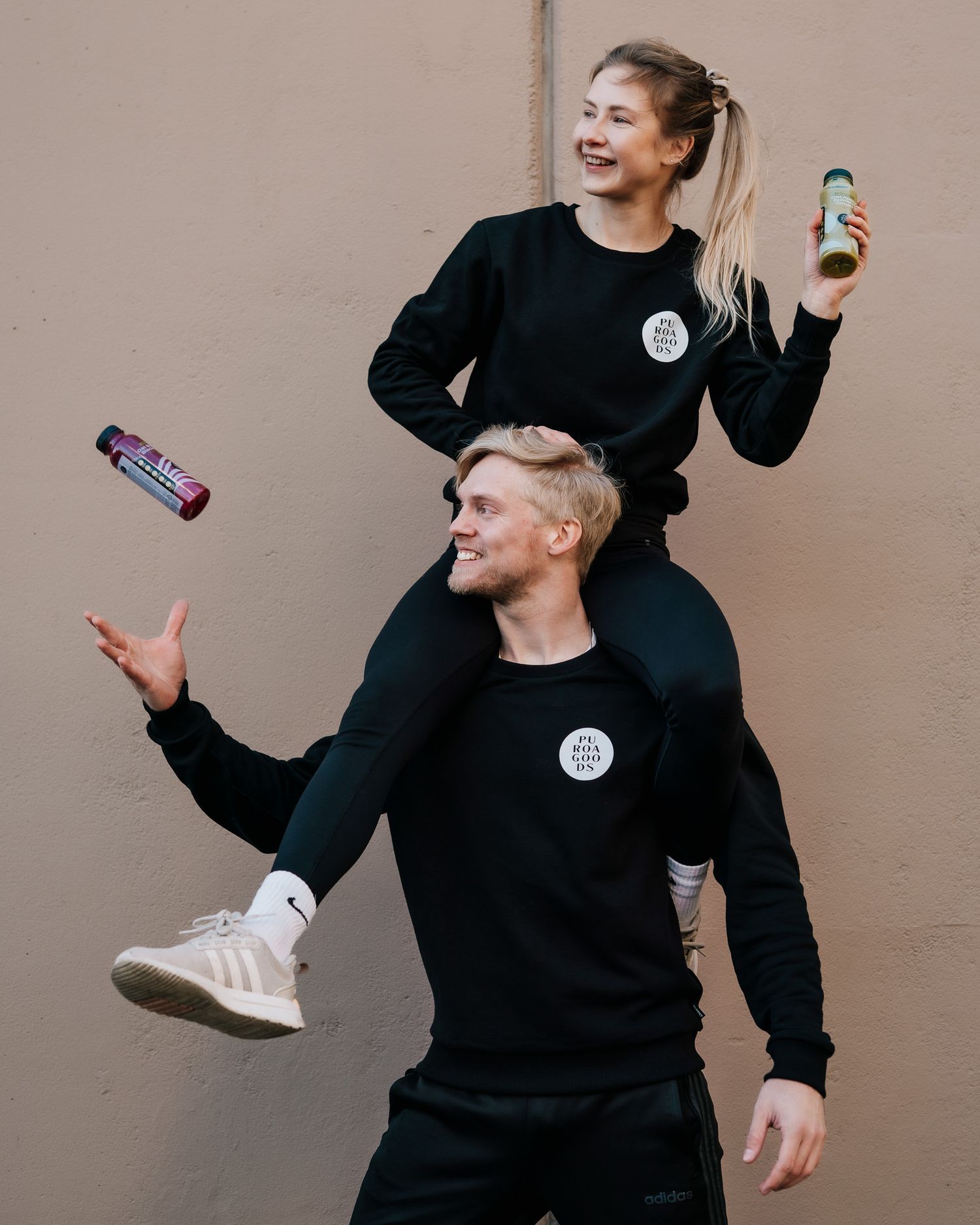 Two people in matching black sweatshirts playfully pose with energy drinks against a beige wall.