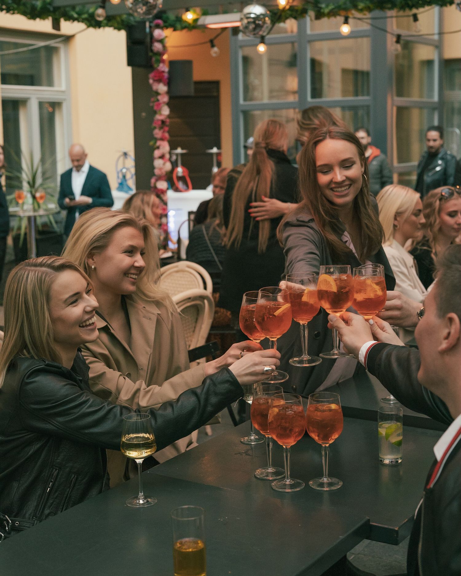 A group of friends raise their glasses in a cheerful toast at an outdoor patio bar with festive decorations.