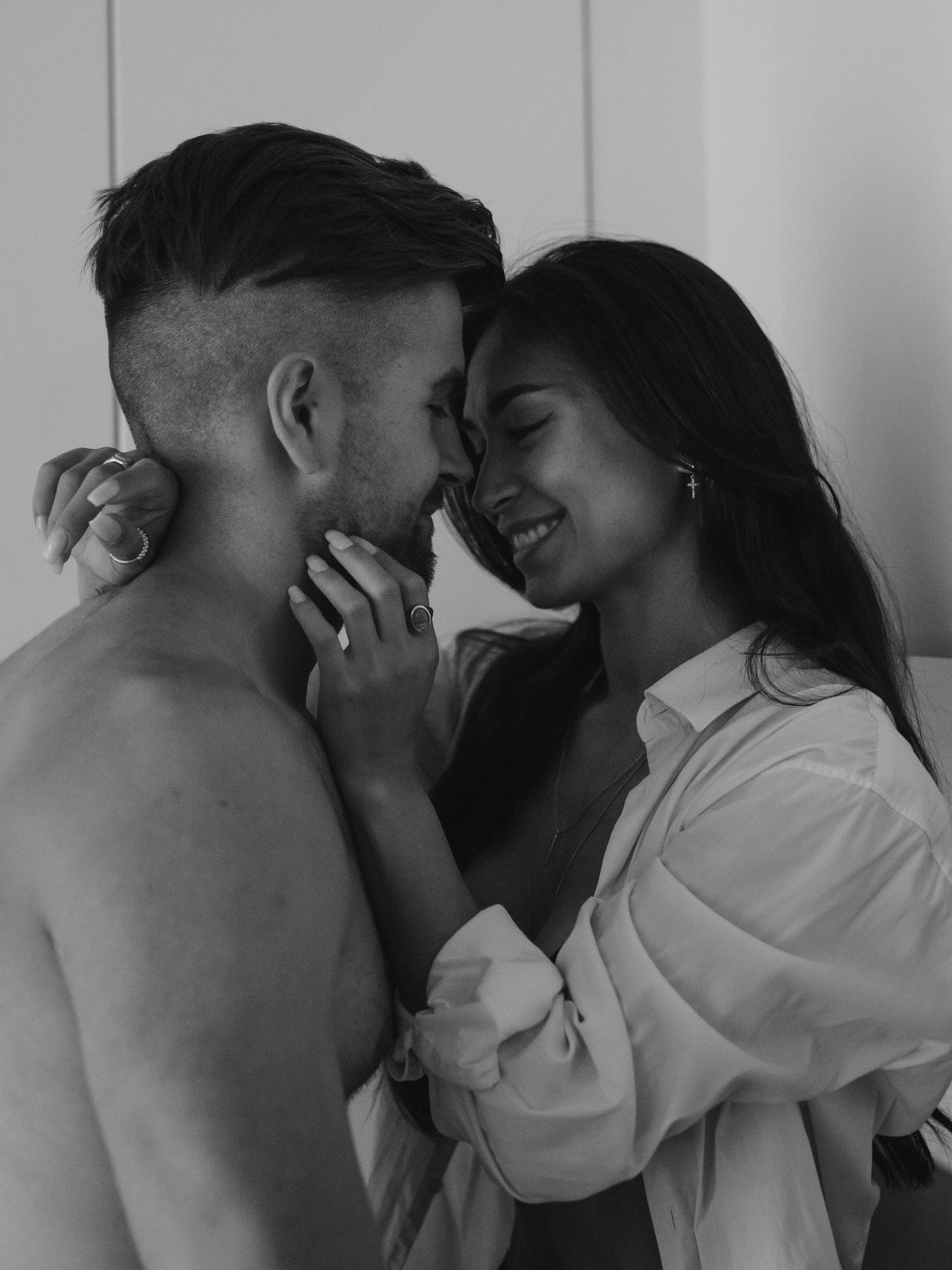 Black and white intimate portrait of a romantic couple sharing a tender close moment against a light colored wall.