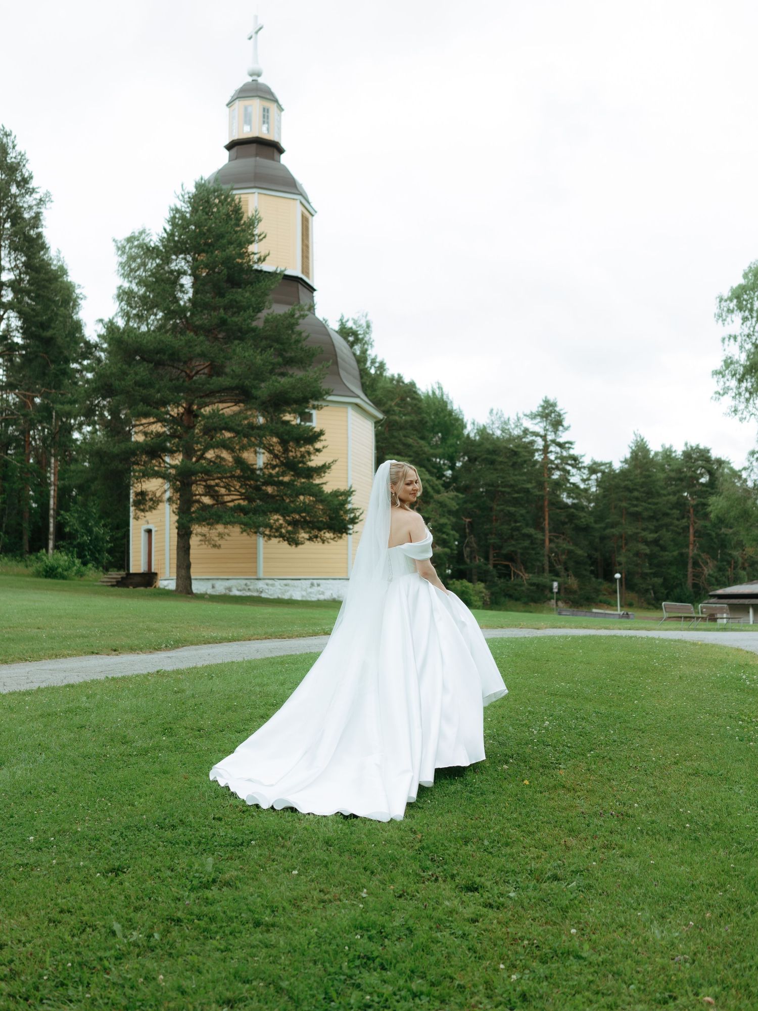 Bride in white gown and veil twirling outside a yellow wooden church surrounded by green trees.