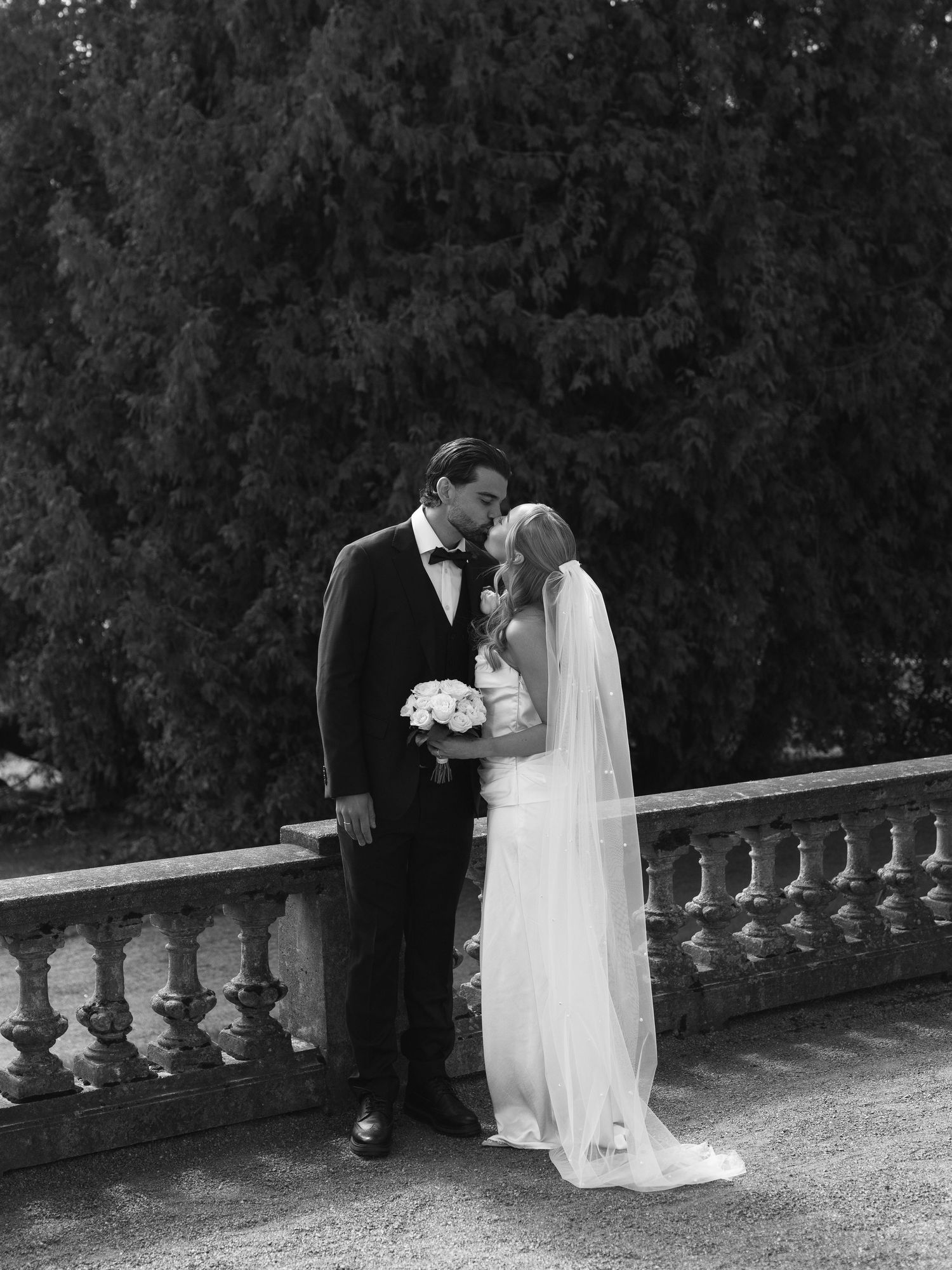 Black and white photo of a bride and groom sharing a kiss on a stone balcony surrounded by tall evergreen trees.
