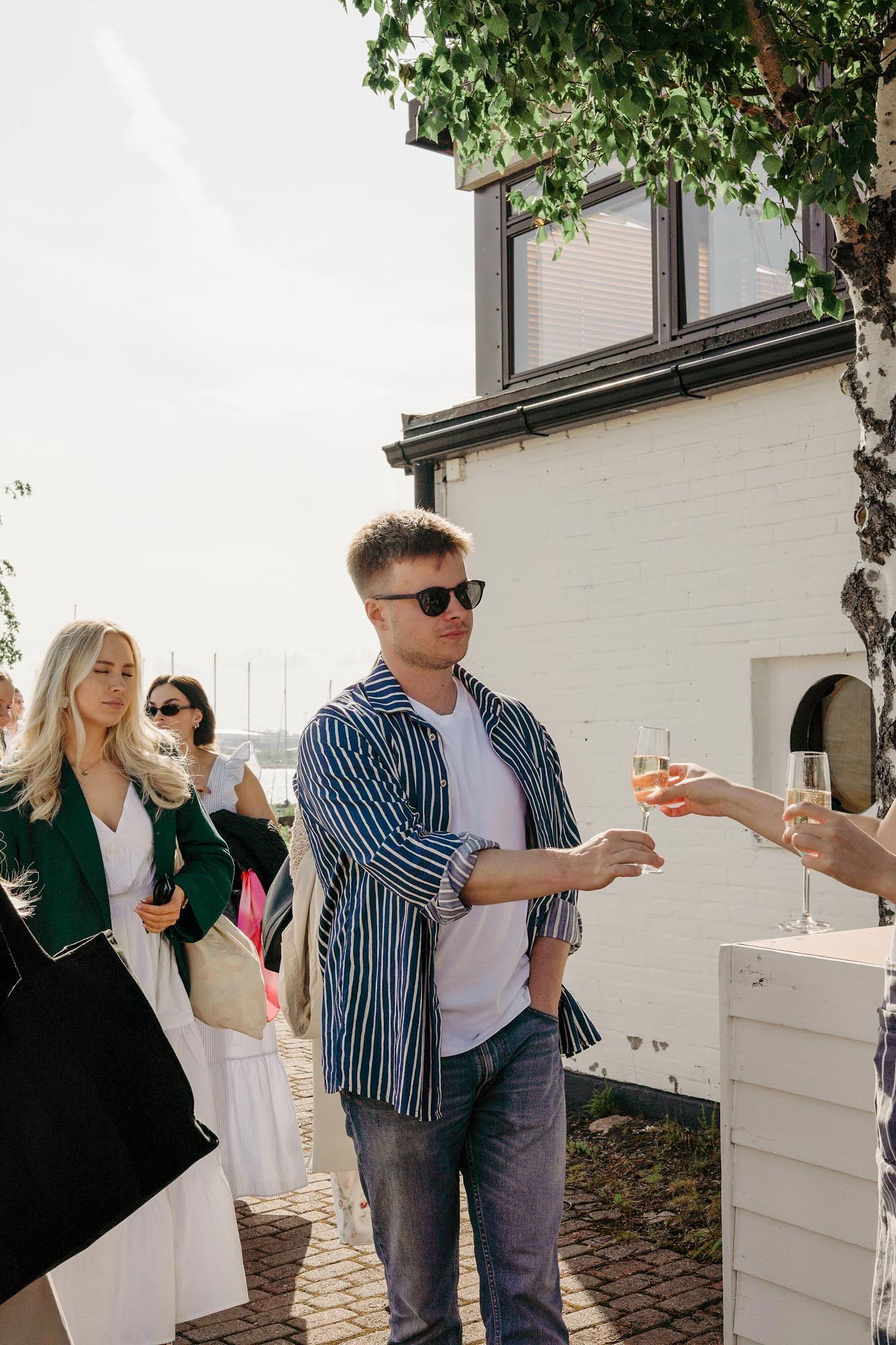 Man in striped jacket and sunglasses receiving champagne glass at outdoor summer social gathering near white building.
