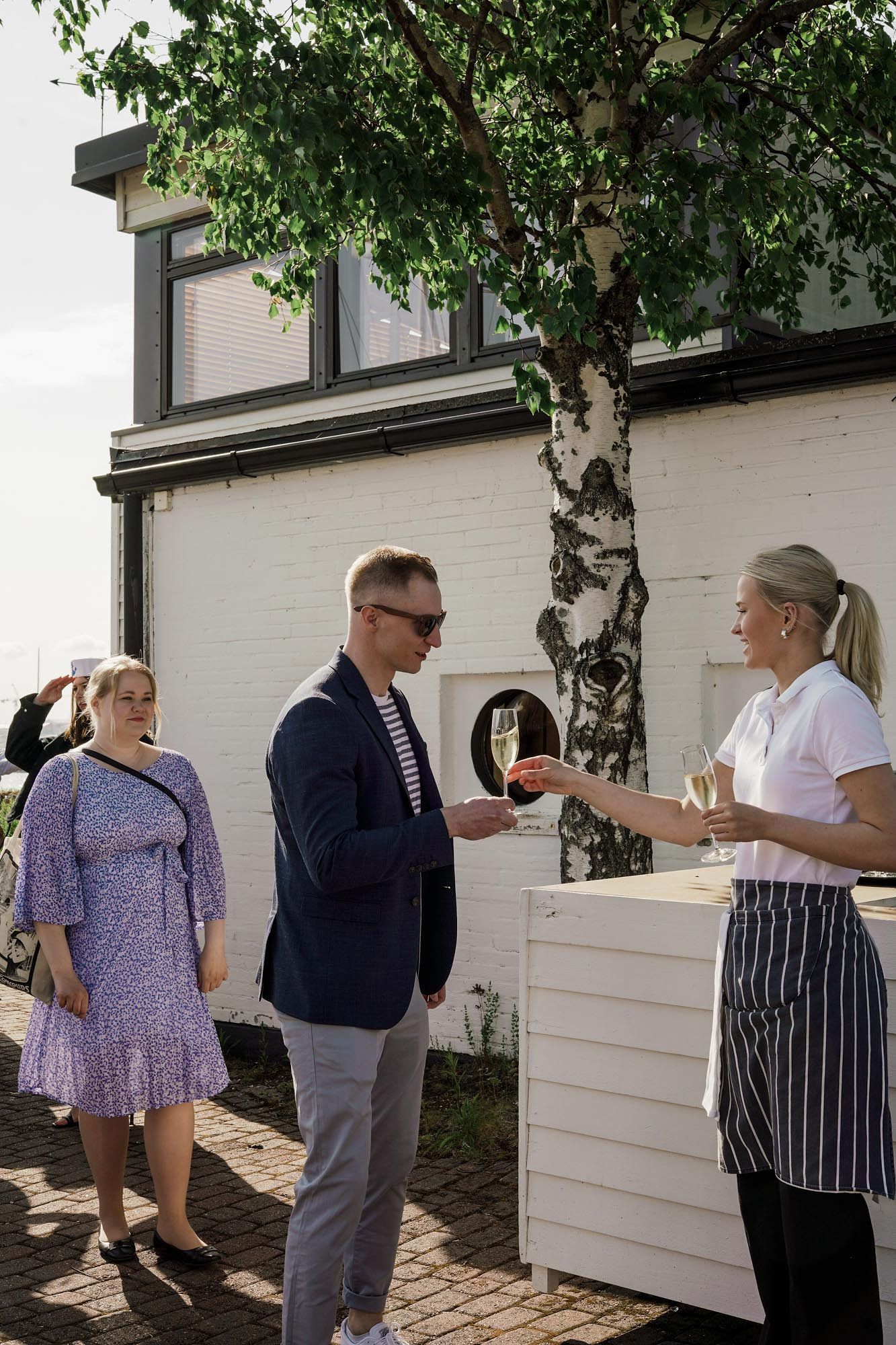 Staff member in apron serves champagne to elegantly dressed guests outside a white brick building on a sunny day.