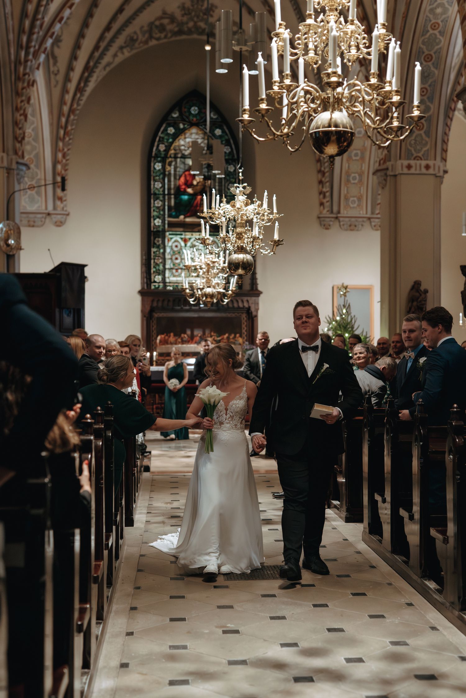 A couple walks down an elegant church aisle under ornate chandeliers during a glamorous wedding ceremony.