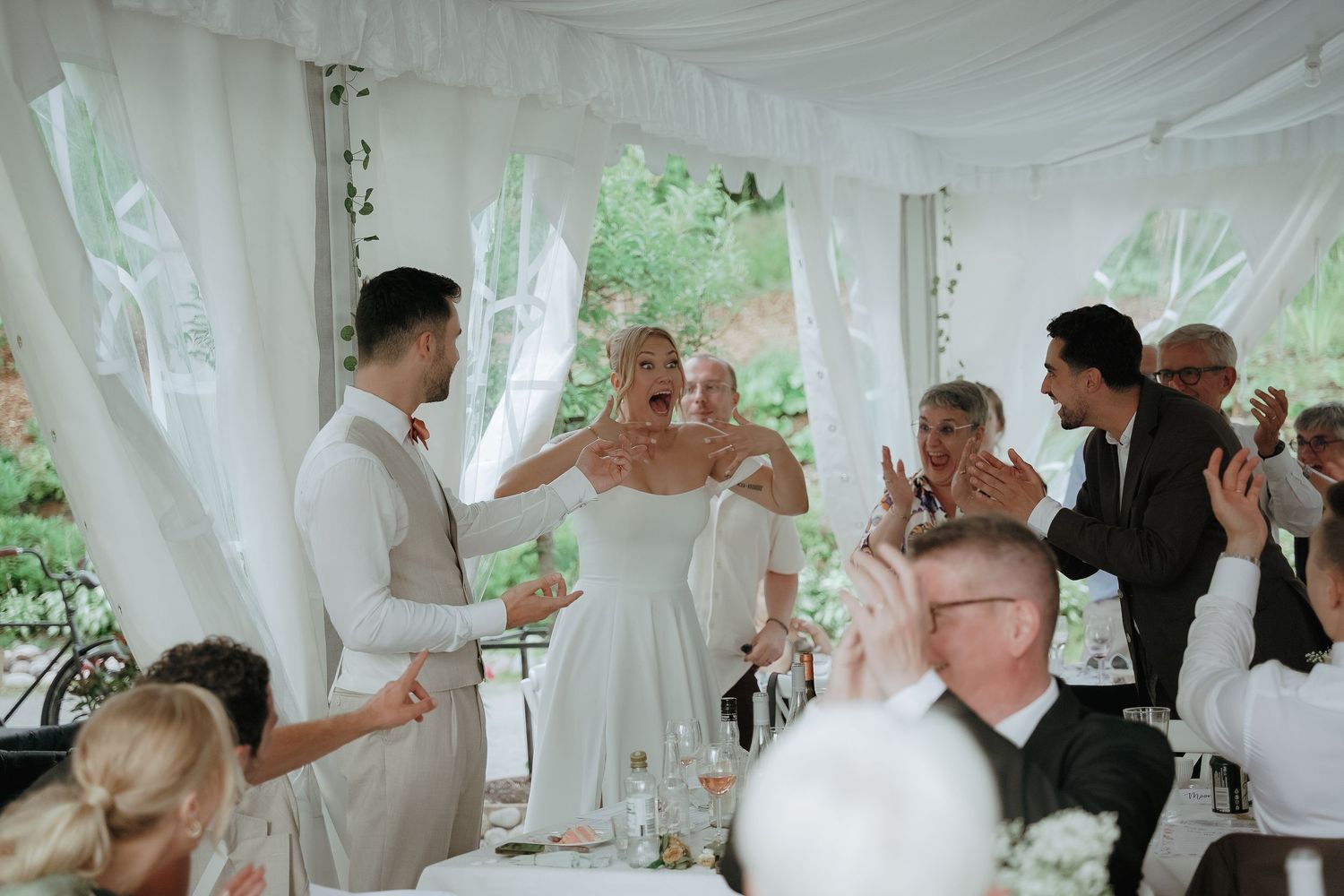 Wedding reception guests dancing and celebrating under white tent with flowing fabric decor.