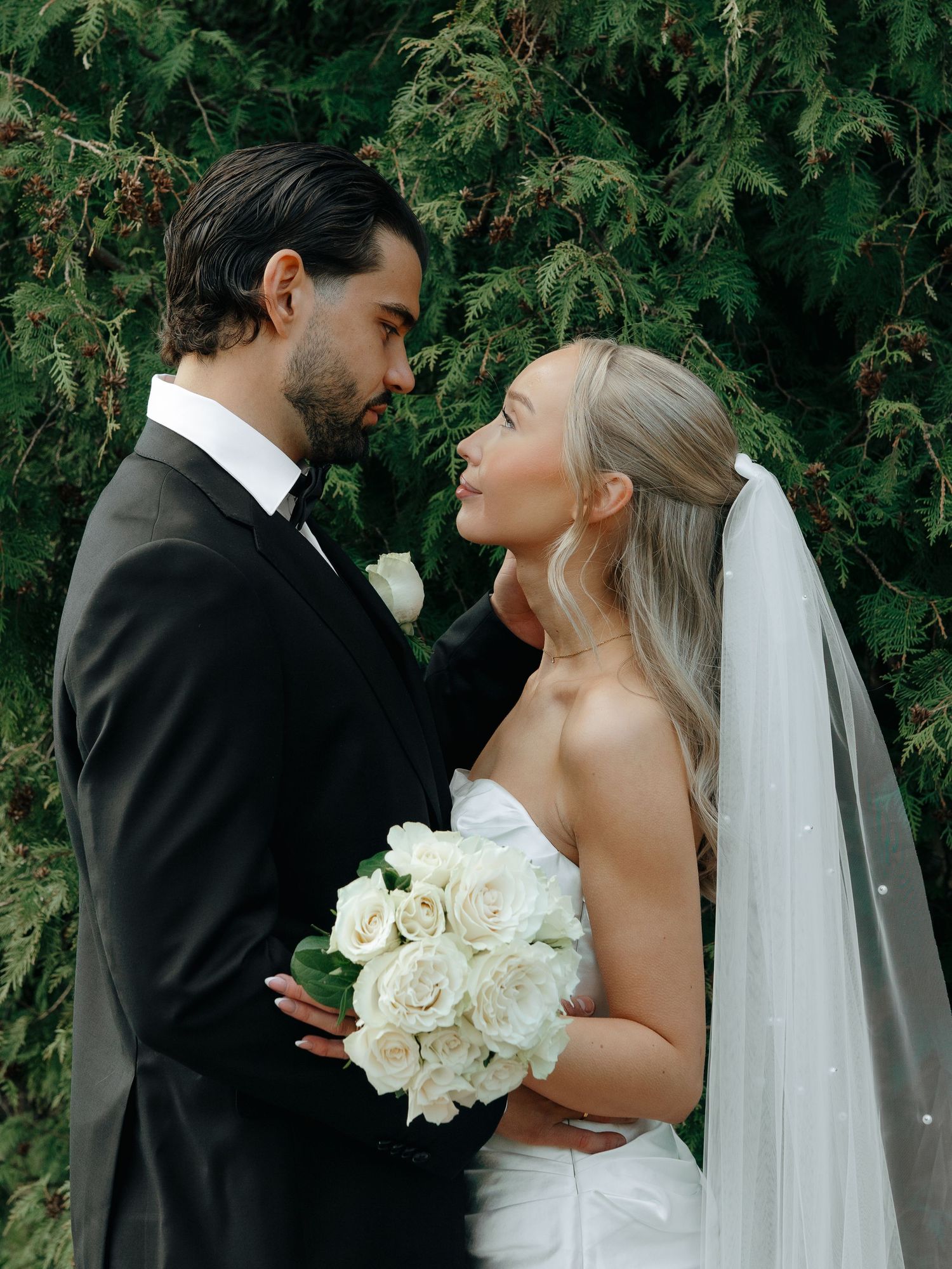 Bride and groom gazing at each other, she holds white roses bouquet, wearing veil and strapless gown, he in black tuxedo.