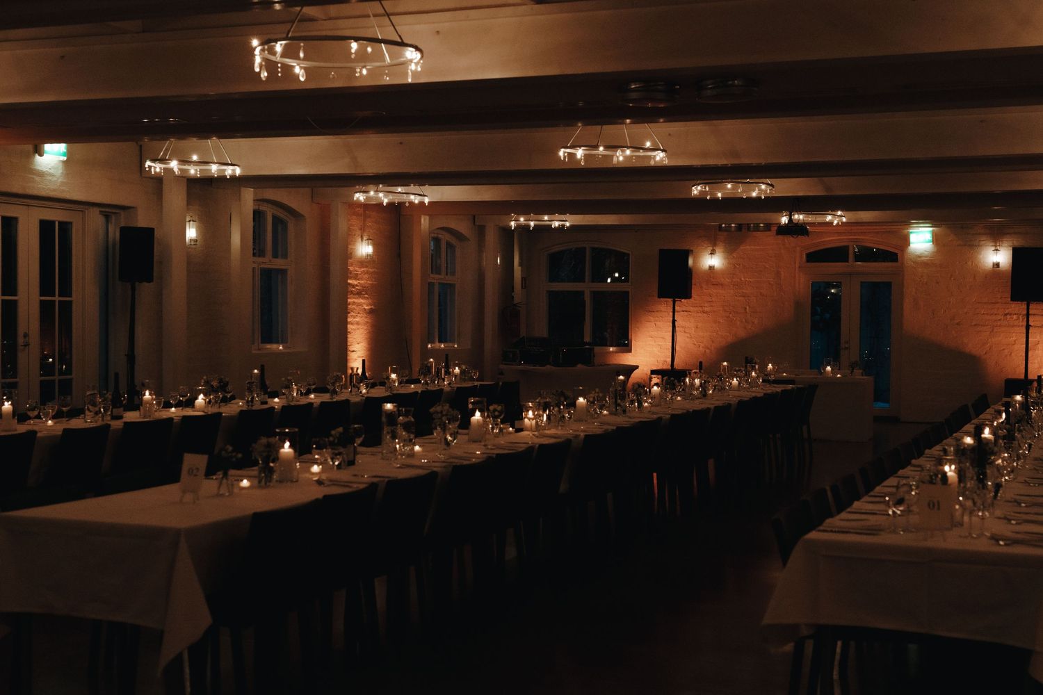 Long banquet tables with white linens and candle centerpieces set in a dimly lit room with warm ambient lighting.