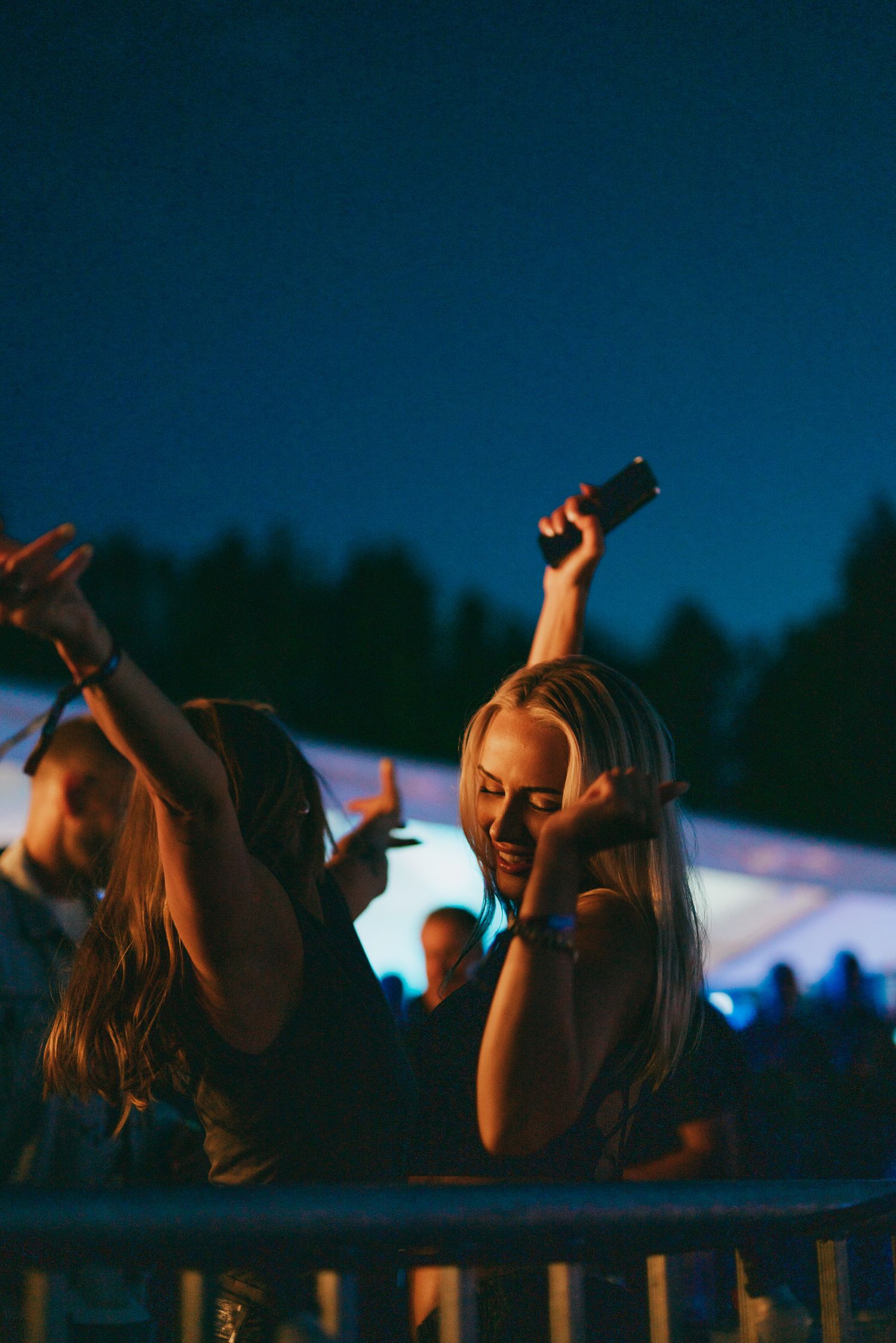 People enjoying a nighttime concert or festival with arms raised and lights in background at outdoor venue.