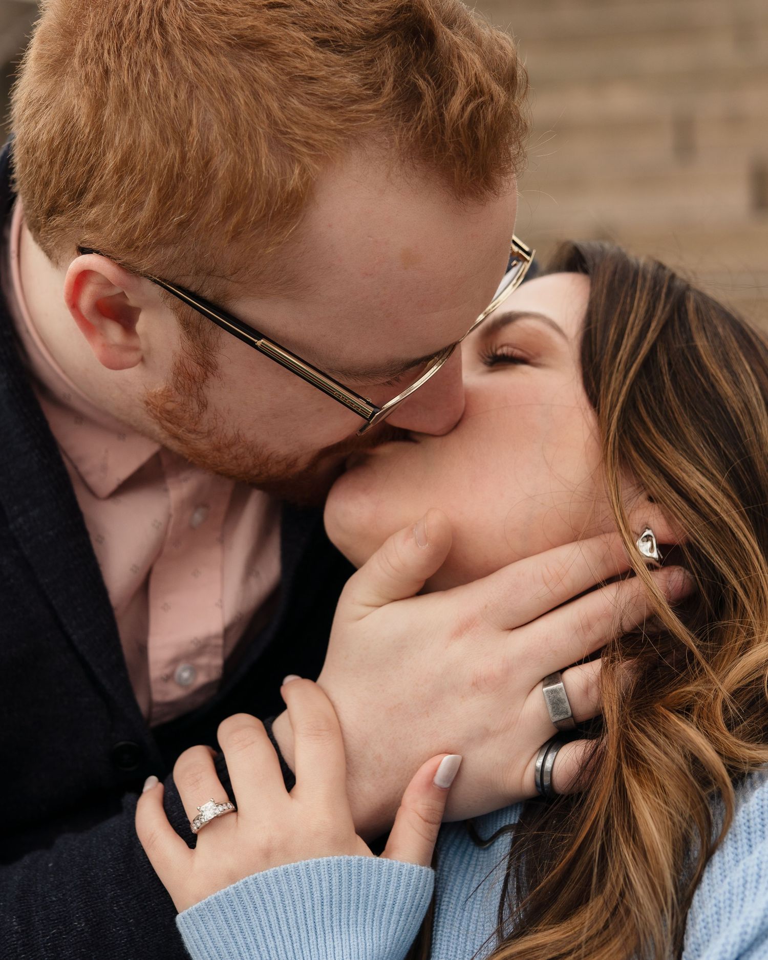A couple sharing a romantic kiss, the man with red hair and glasses, both wearing rings, in a close embrace.