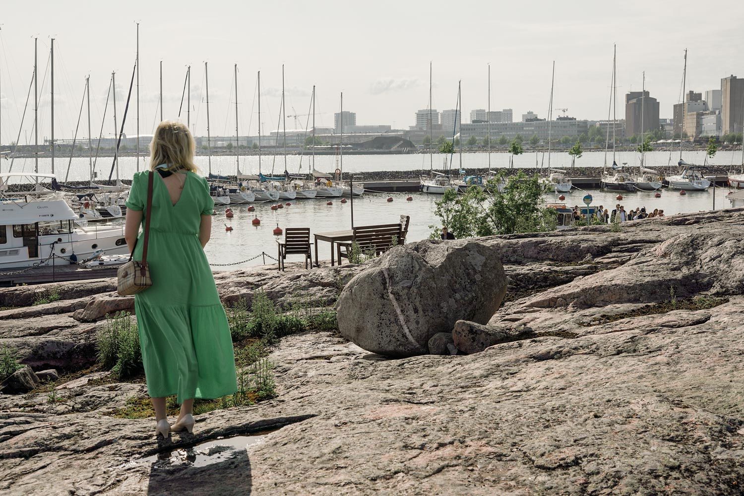 Woman in green dress standing on rocky shoreline overlooking a marina with sailboats and city skyline.