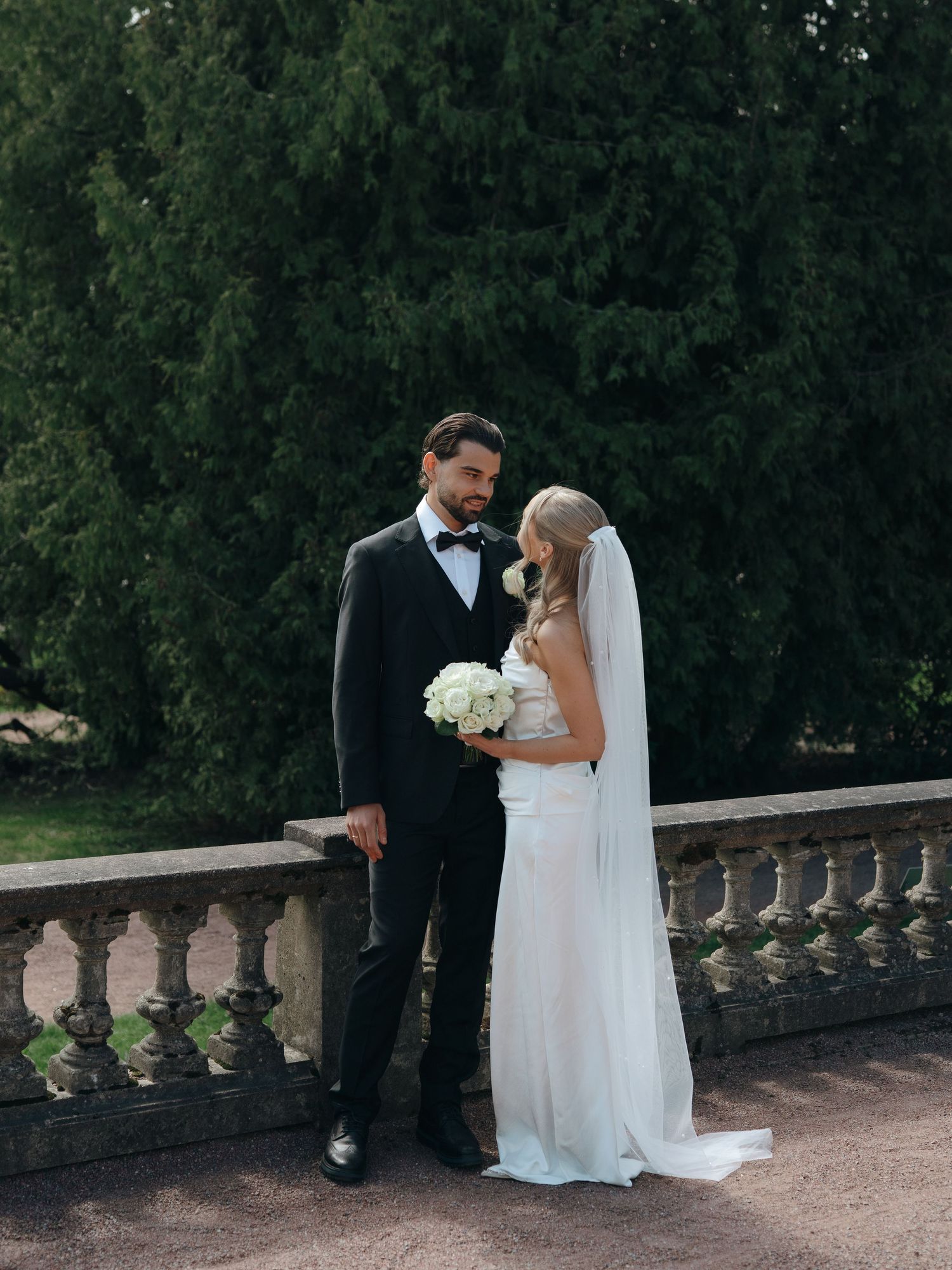 Elegant bride and groom posing on a stone balustrade terrace, bride holding white bouquet, groom in black tuxedo.
