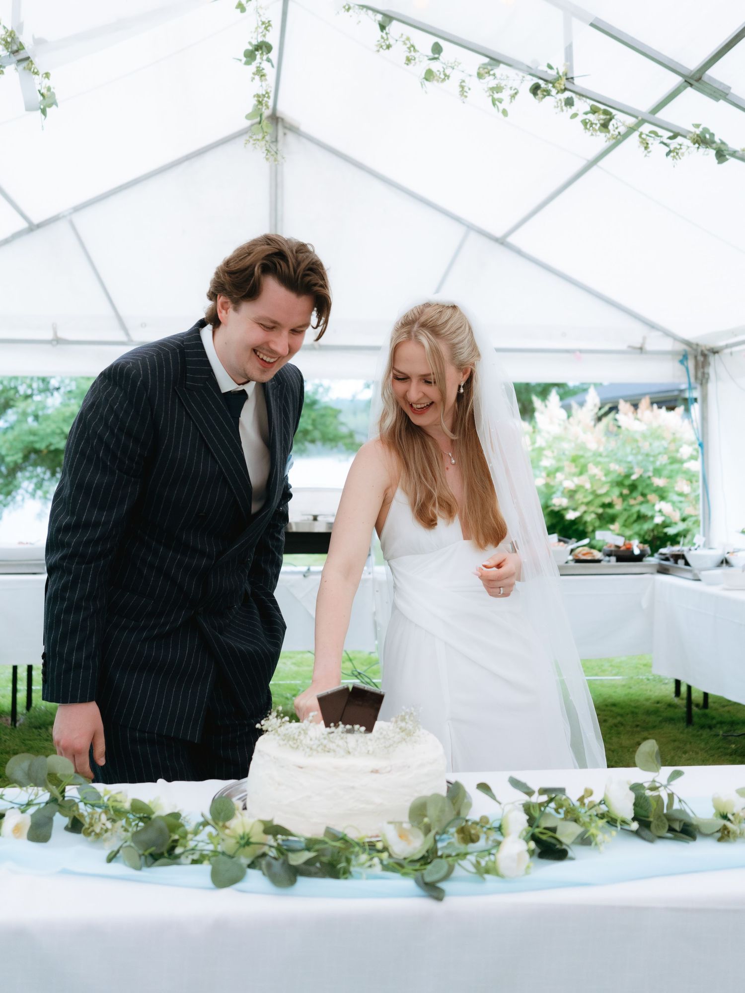Bride and groom cutting white wedding cake under marquee tent decorated with eucalyptus greenery.