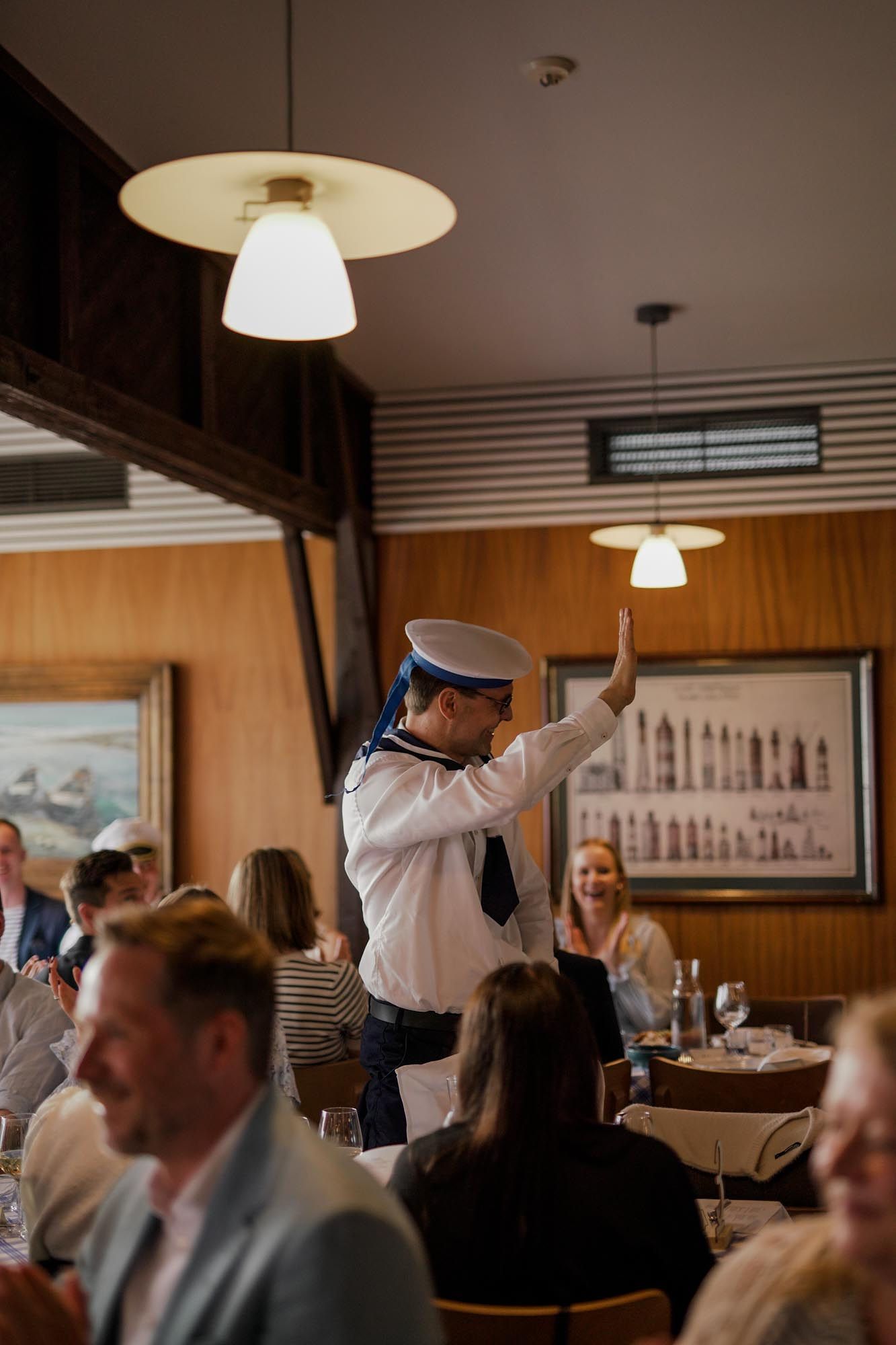 A waiter in a sailor hat gestures to diners in a warm, wood-paneled restaurant with pendant lights.