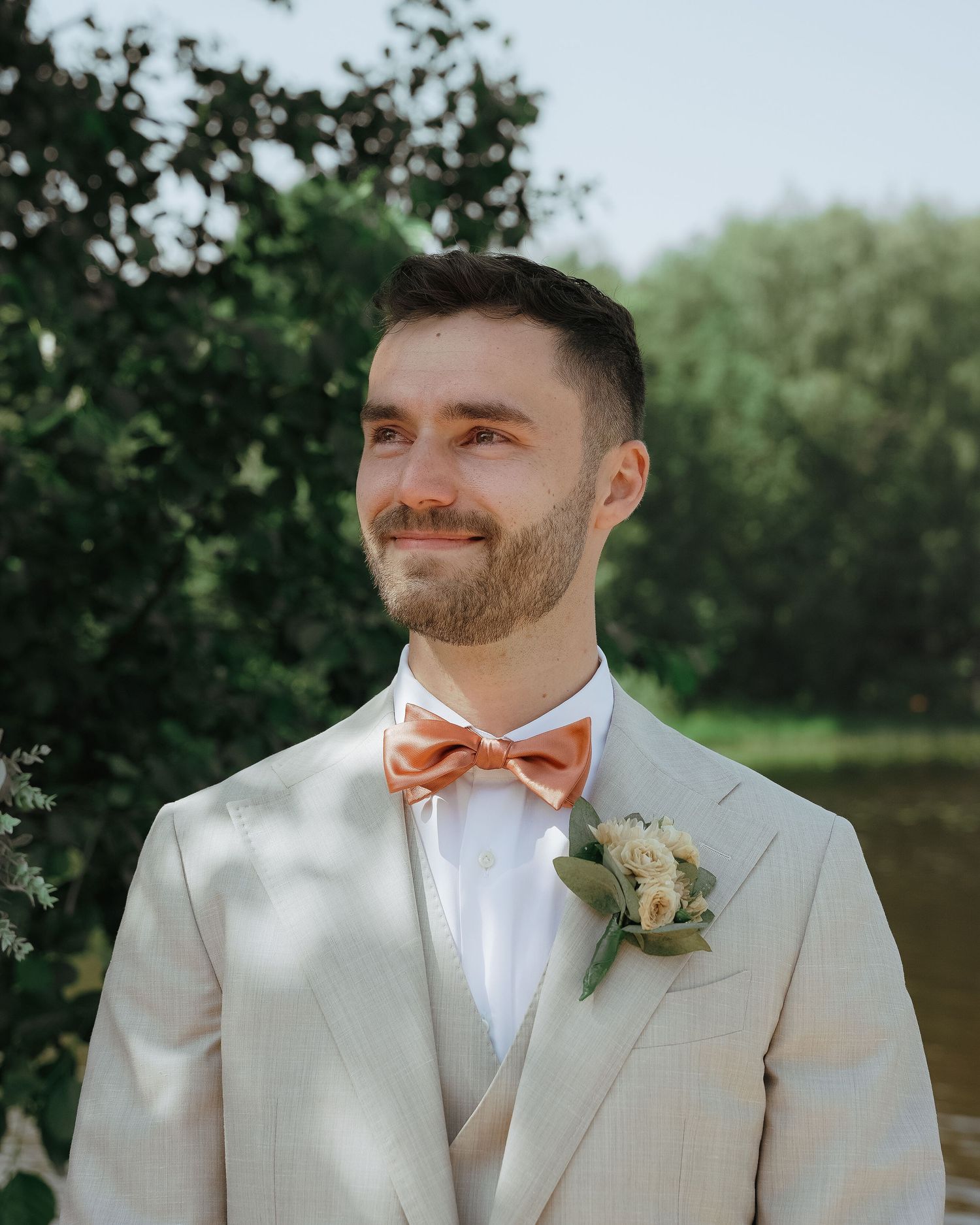 A series of wedding portraits showing a groom in a light beige suit with peach bow tie outdoors with greenery in background.