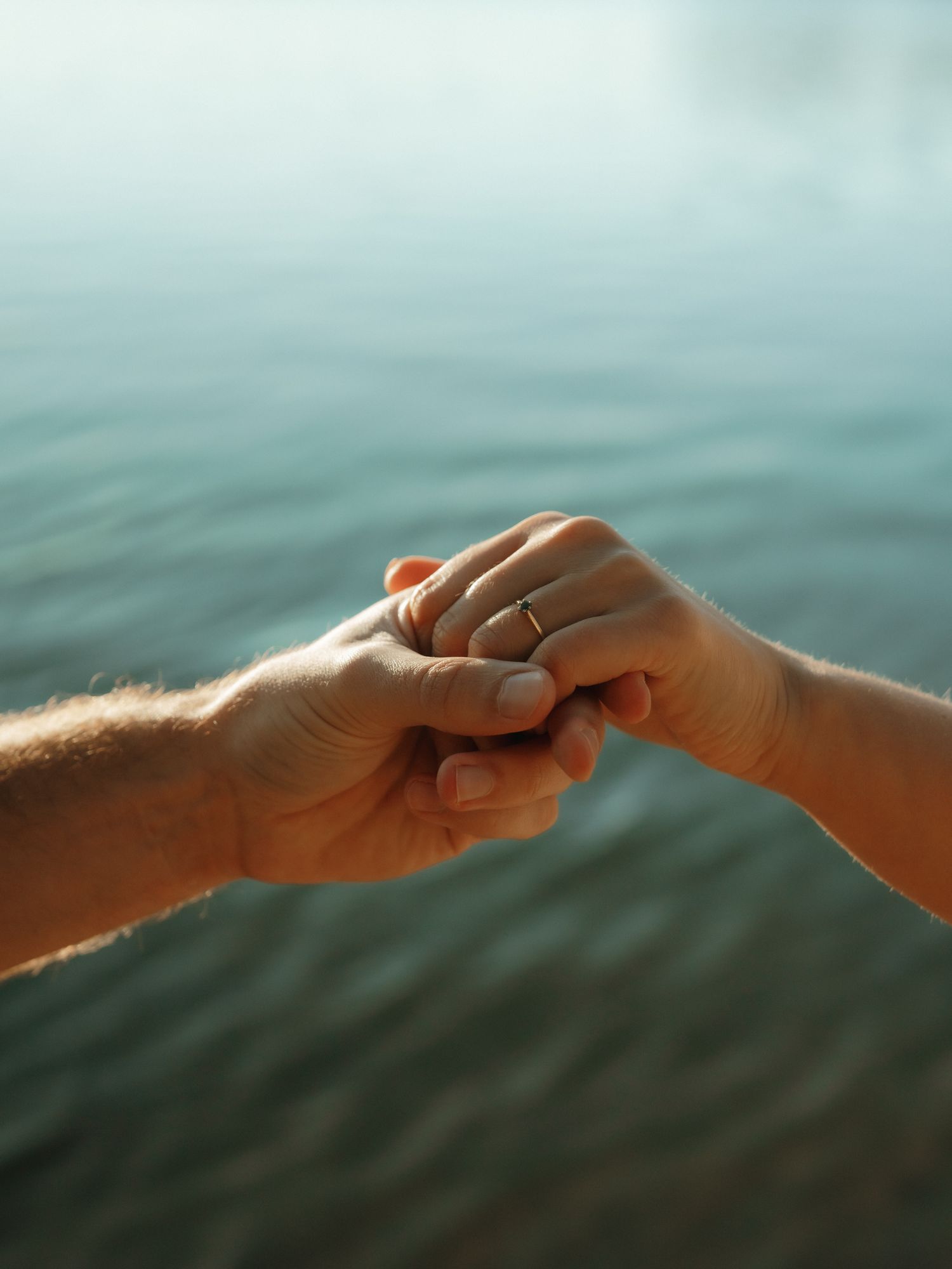 Two hands gently clasping together against a blurred teal water background, with one hand wearing a ring.