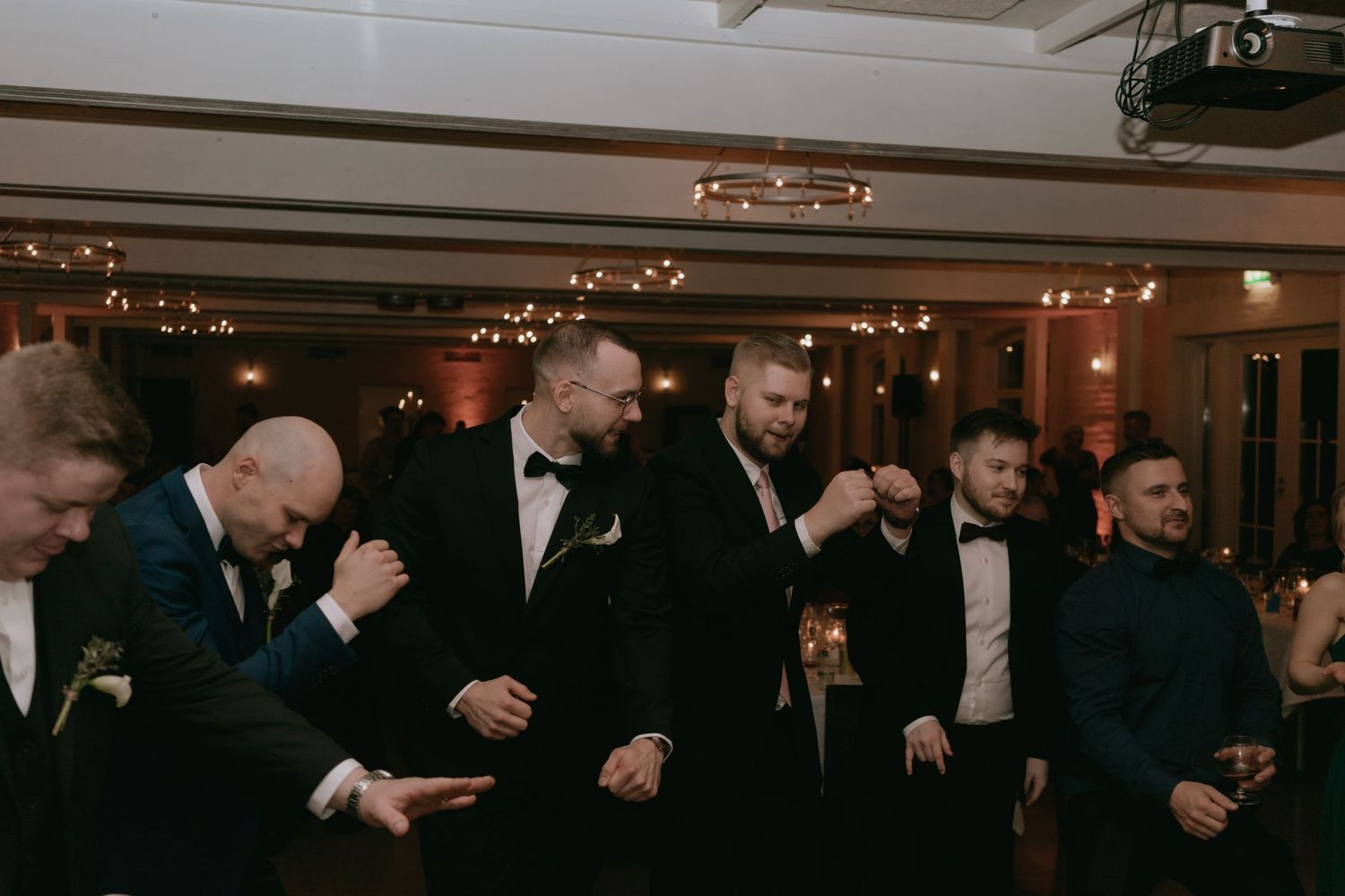 A group of groomsmen in black tuxedos dance and laugh together at a dimly lit wedding reception.