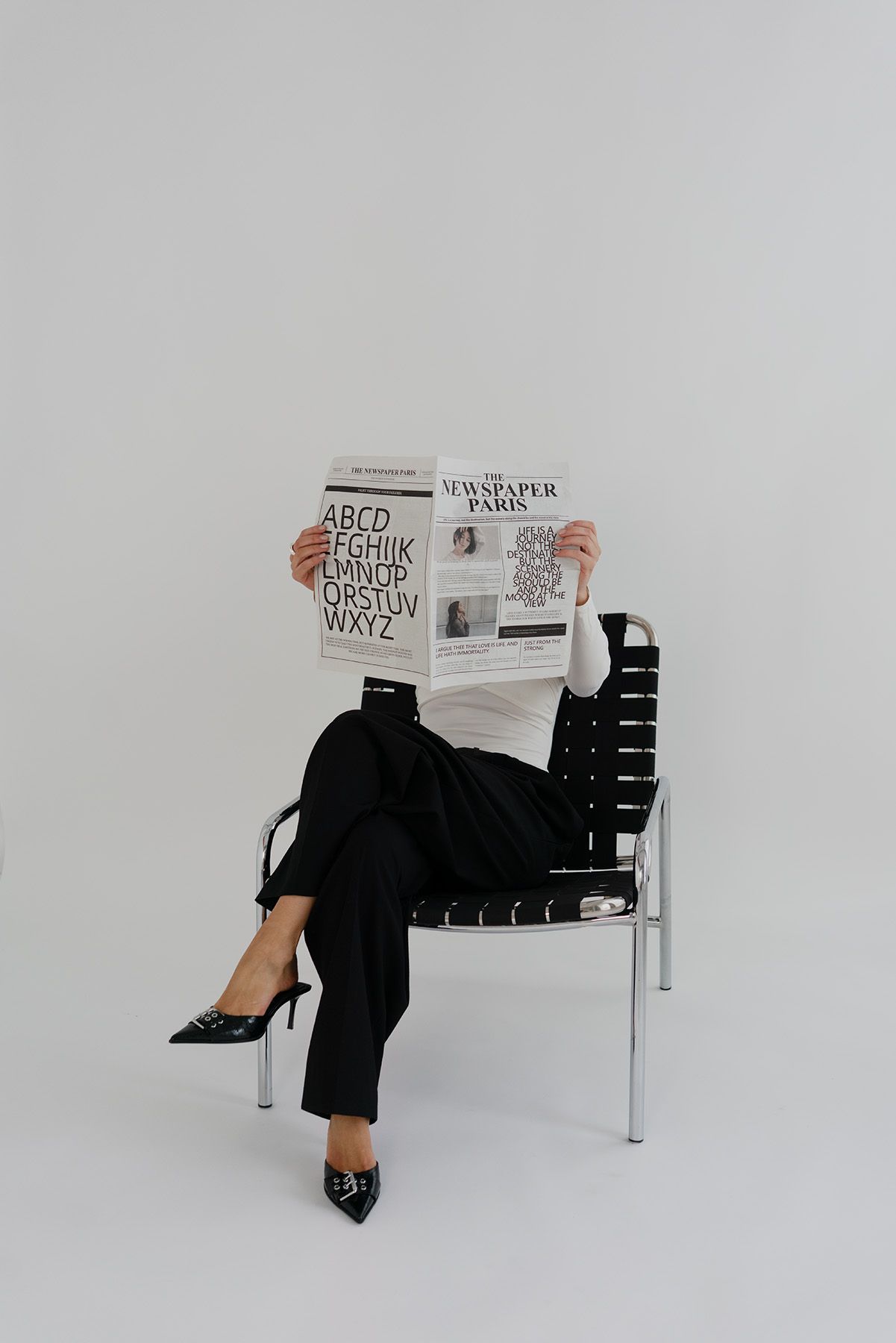 A person in black attire reads a newspaper while seated in a modern woven chair.