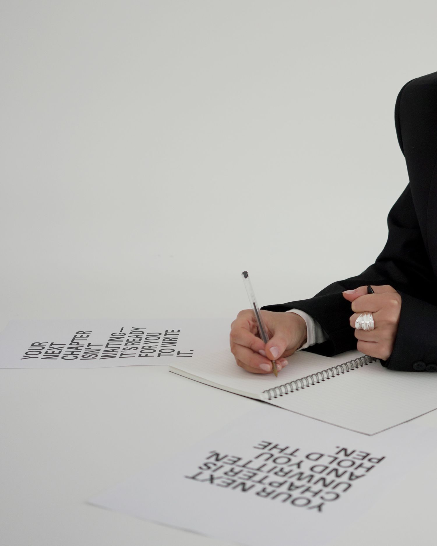 Someone in black clothing writes in a notebook while text appears to float above the white paper in an artistic composition.