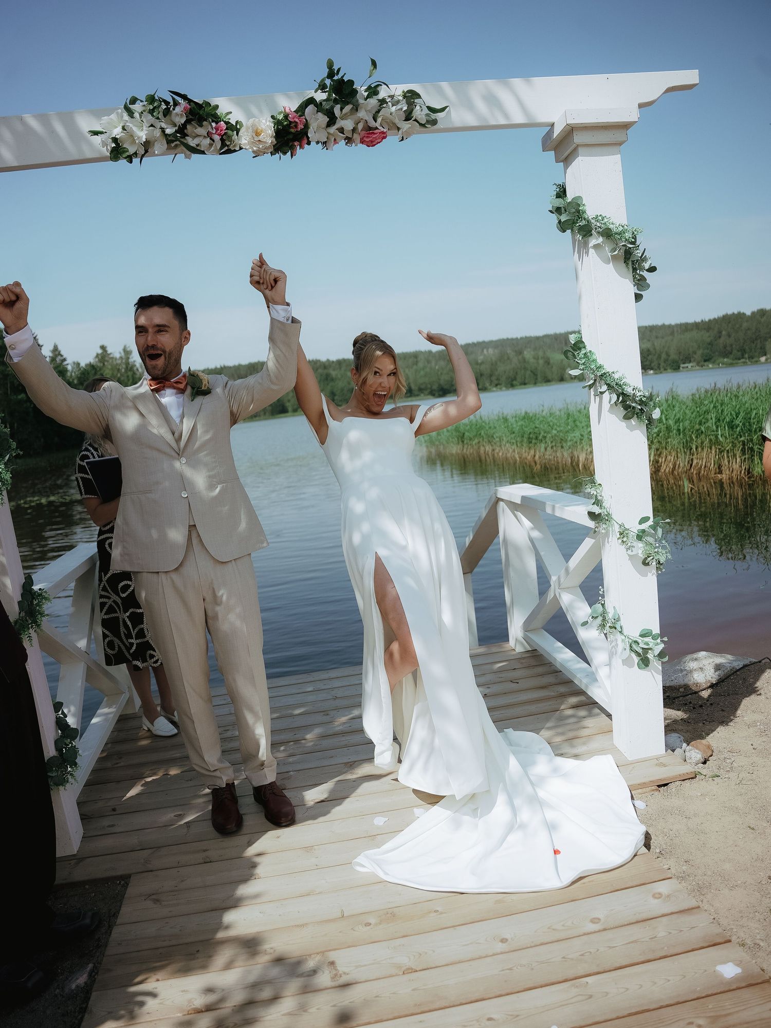 A couple celebrates joyously under a floral arch on a waterfront dock during their wedding ceremony.