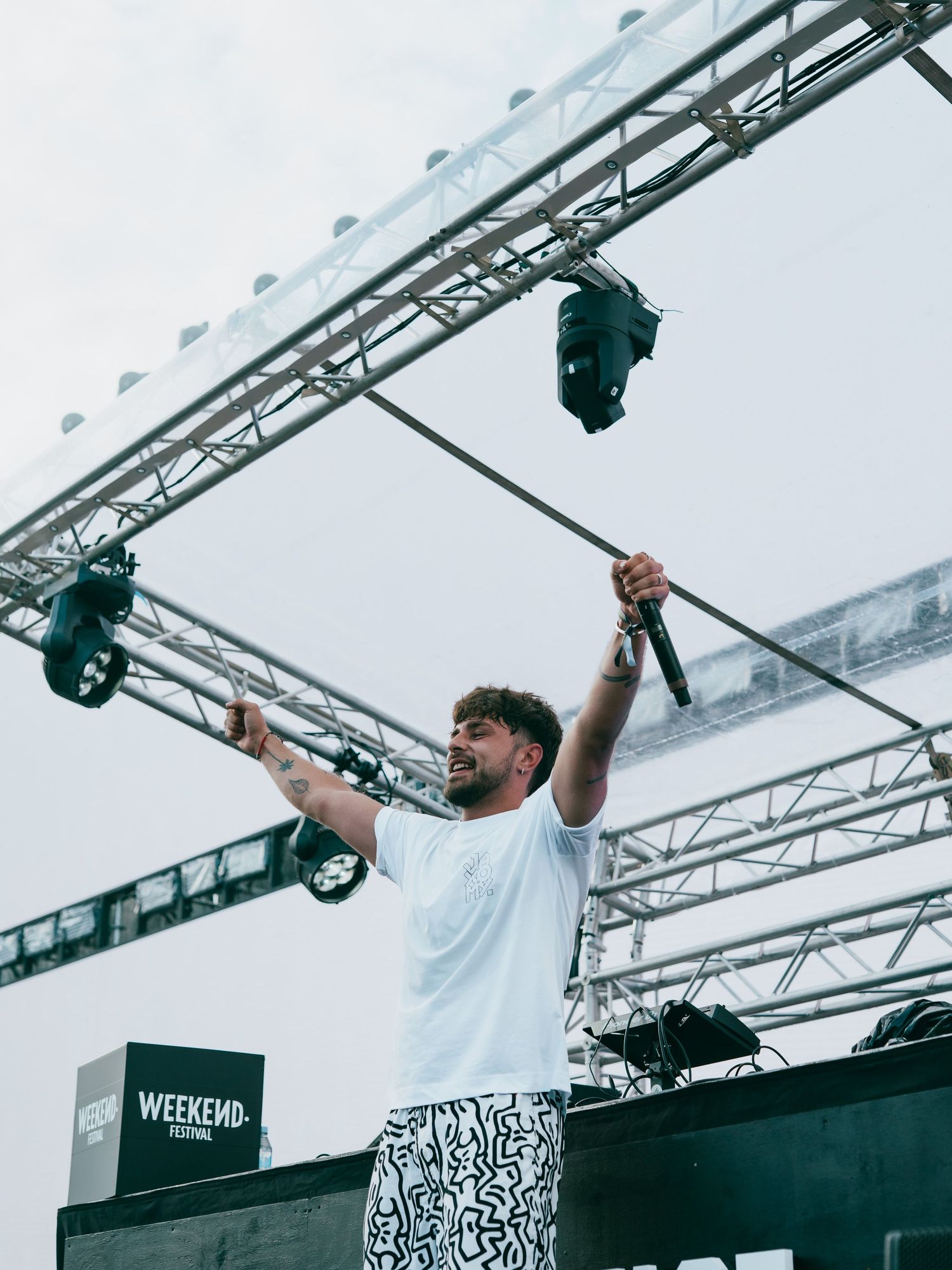 A performer in a white t-shirt with patterned pants raises their arms triumphantly on stage under lighting equipment at an event.