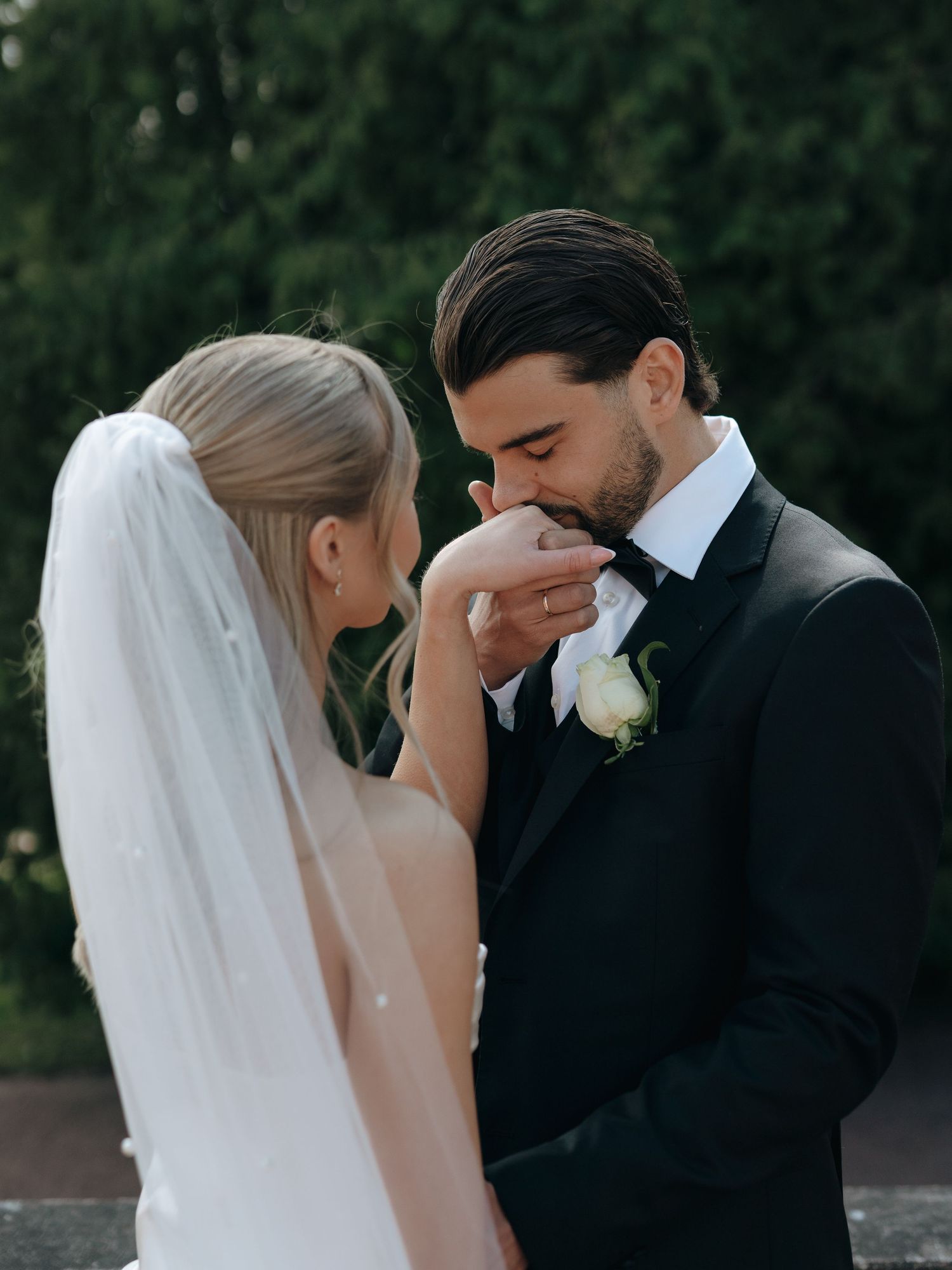 Groom in black tuxedo kissing bride's hand while she wears a white gown and veil at outdoor wedding.
